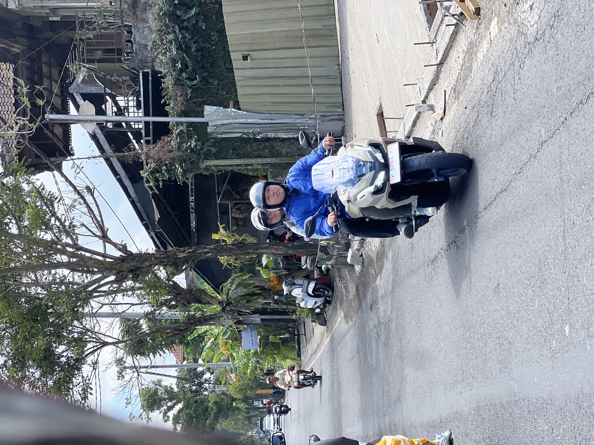 The image depicts a street scene in Ubud District, Indonesia, likely on a sunny day. In the foreground, two individuals are riding a modern scooter. The person in the front, wearing a blue jacket and a helmet, is driving, while the person behind, also wearing a helmet, is the passenger. The scooter has a prominent windshield and a license plate displaying "DK 5703 XY". To the left of the scooter, another scooter is parked, and further down the street, several other motorcycles and cars are visible. The street is lined with trees, some of which are large and leafy, and buildings with traditional Indonesian architecture. One building in the background appears to be under construction or renovation, with exposed structural elements. The environment suggests a typical urban or semi-urban setting in Indonesia, with a focus on the use of motorcycles as a primary mode of transportation.