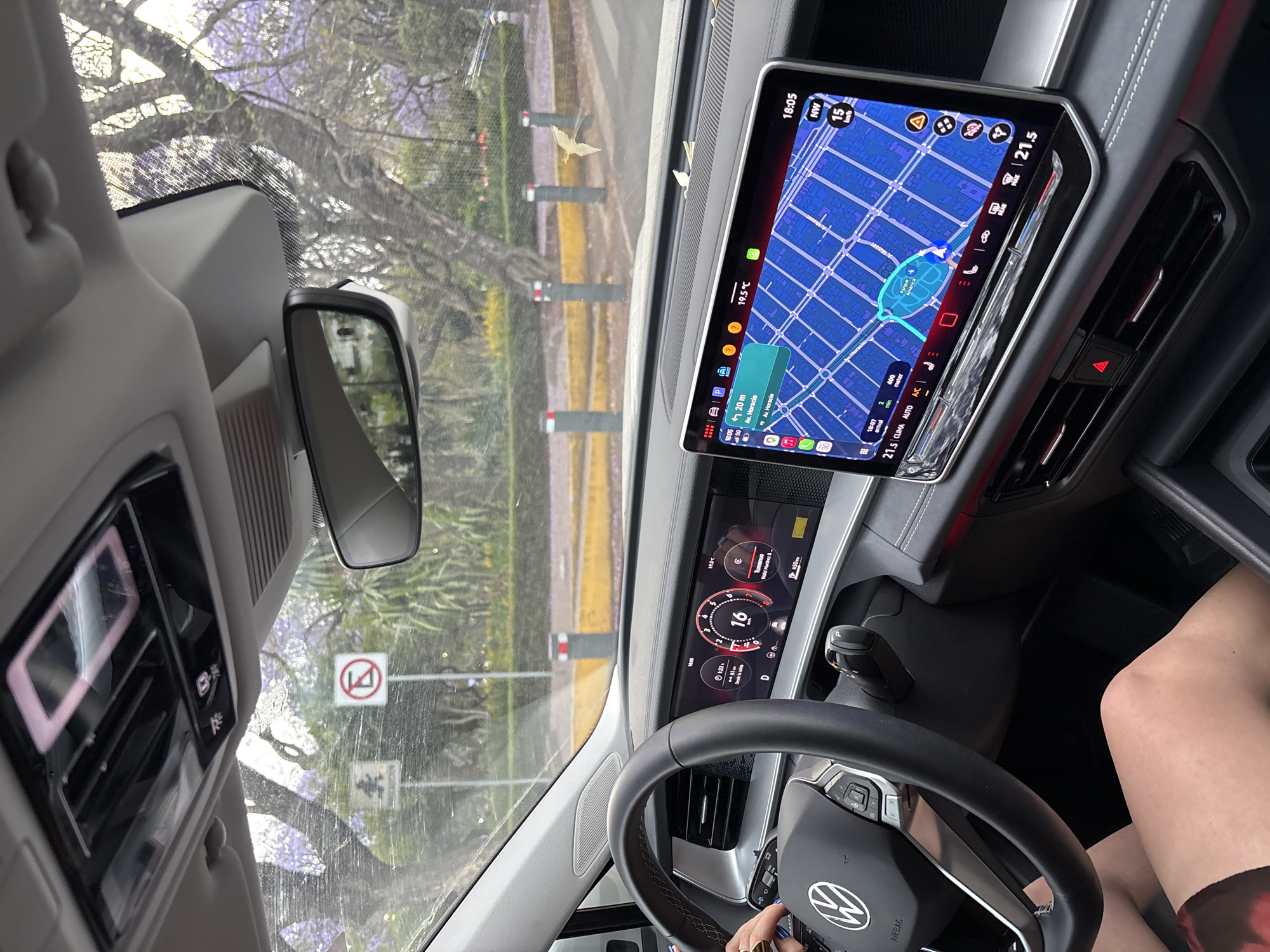 Here is a forensic description of the image:
The image is taken from inside a Volkswagen car. The dashboard, steering wheel, and infotainment system are visible. The infotainment system displays a map with a route set to Av. Horacio. The car is driving at 16 km/h.
Outside the car, there is a street with trees. A traffic sign can be seen through the windshield.
The time on the infotainment system is 18:05, and the temperature is 19.5°C.