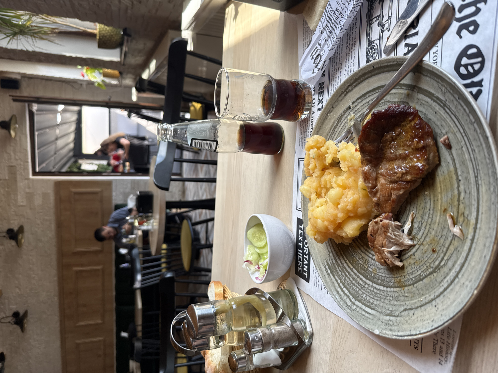 The image depicts a close-up view of a table in what appears to be a restaurant. In the foreground, a plate holds a meal consisting of mashed potatoes and a piece of meat, with a fork resting on the side. To the left of the plate are salt and pepper shakers and a bottle of oil and vinegar. A small bowl of salad, a glass of dark beverage, and a bottle of cola are also visible on the table. The background shows other tables, chairs, and patrons, suggesting a lively dining atmosphere. The restaurant's interior features wooden panels, brick walls, and hanging lamps. The overall impression is one of a casual dining experience.
