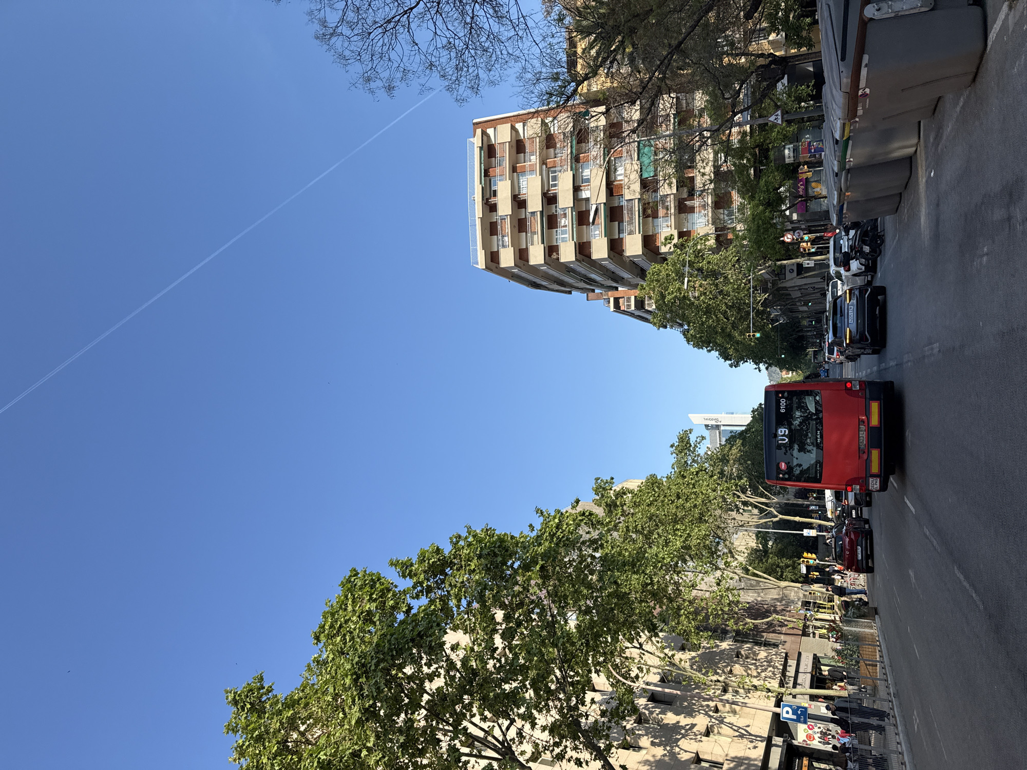 The image captures a street scene in Barcelona, Spain, showcasing urban life on a sunny day. A red city bus, marked with "U9" and "6100", occupies the right lane of the street, followed by several cars. On the right sidewalk, pedestrians walk and stand near storefronts. A building with several apartments is to the left of the bus. Overhead are a few bare branches and the blue sky. There is a single contrail in the sky.
