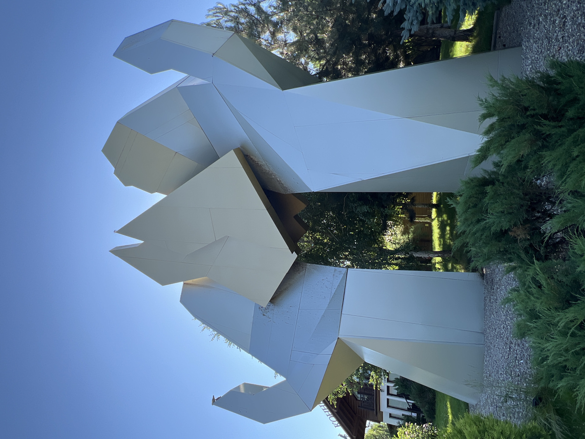 The image captures a large, abstract metallic sculpture with sharp, geometric angles. The sculpture is silver and gold in color and appears to be composed of several large, angular panels. It is situated outdoors in a well-maintained green space with trees and shrubs. To the left of the sculpture, a portion of a wooden building with a white facade and windows is visible, with a sign that reads "КОНН" above the entrance. The background features a clear blue sky, sunlight, and a variety of trees, including a tall evergreen tree on the right side. The ground around the sculpture is covered with small gray gravel, and lush green bushes are in the foreground. The location is identified as Plastunivska, Russia. There are no people visible in the image.