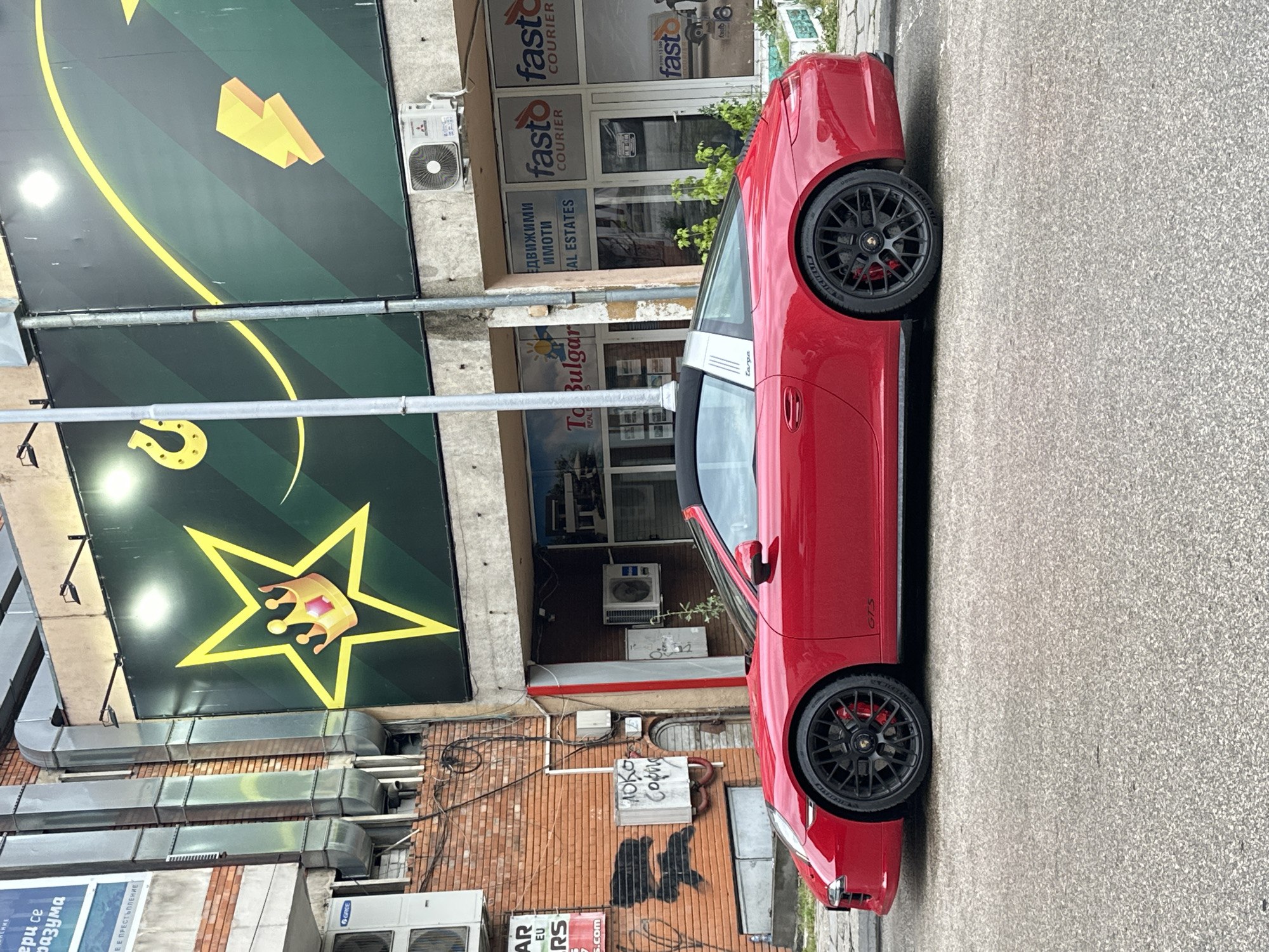 The image shows a red Porsche 911 Targa 4 GTS parked on a street in Veliko Tarnovo, Bulgaria. The car is facing right, with its black soft-top deployed. The background features a brick building with exposed pipes and air conditioning units, and a large green billboard with a yellow star and crown graphic. Adjacent to the billboard are storefronts for businesses including "Fasto Courier" and "To Bulgarian Real Estates". The street is paved with asphalt. The overall impression is an urban scene with a focus on the sports car.