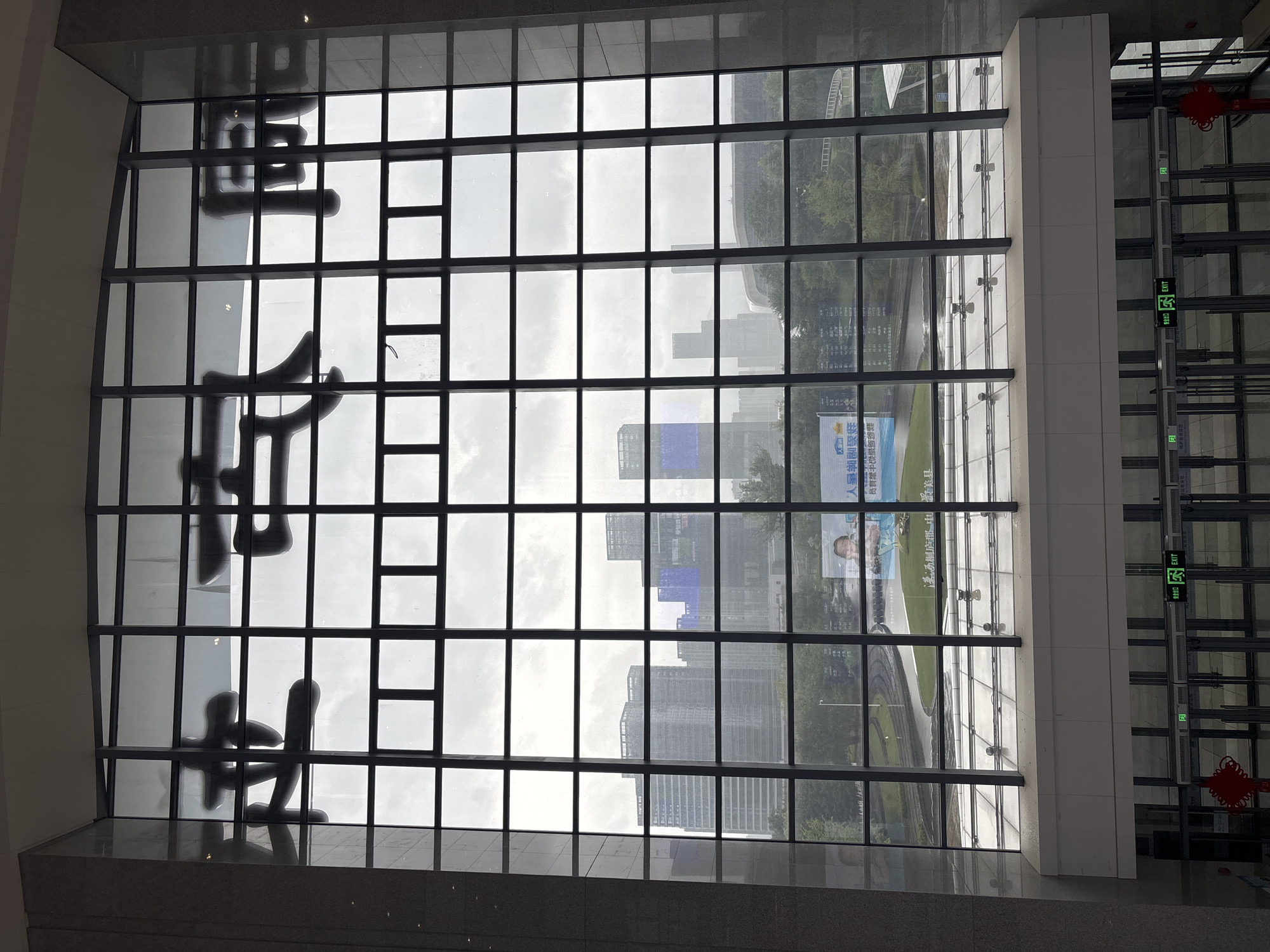 The image shows a large window with dark-colored framing, divided into a grid of squares. On the glass, large, stylized Chinese characters are visible, appearing to be black or dark grey. The characters are: 德 (De), 兴 (Xing), 伟 (Wei), 业 (Ye). Beyond the window, a cityscape is visible under an overcast sky. Several tall buildings, some appearing modern with glass facades and others with more traditional architecture, are prominent. Green trees and a road or pathway are also discernible. A large billboard or advertisement featuring a person's face is visible on the right side of the scene, also beyond the window. The bottom of the image shows a section of a building interior with a white wall and glass doors, some of which are marked with "EXIT" signs. Red decorative knots are visible hanging near the doors. The scene appears to be in Tonglu County, China, likely a public building such as a government center or transportation hub, given the signage and architecture.