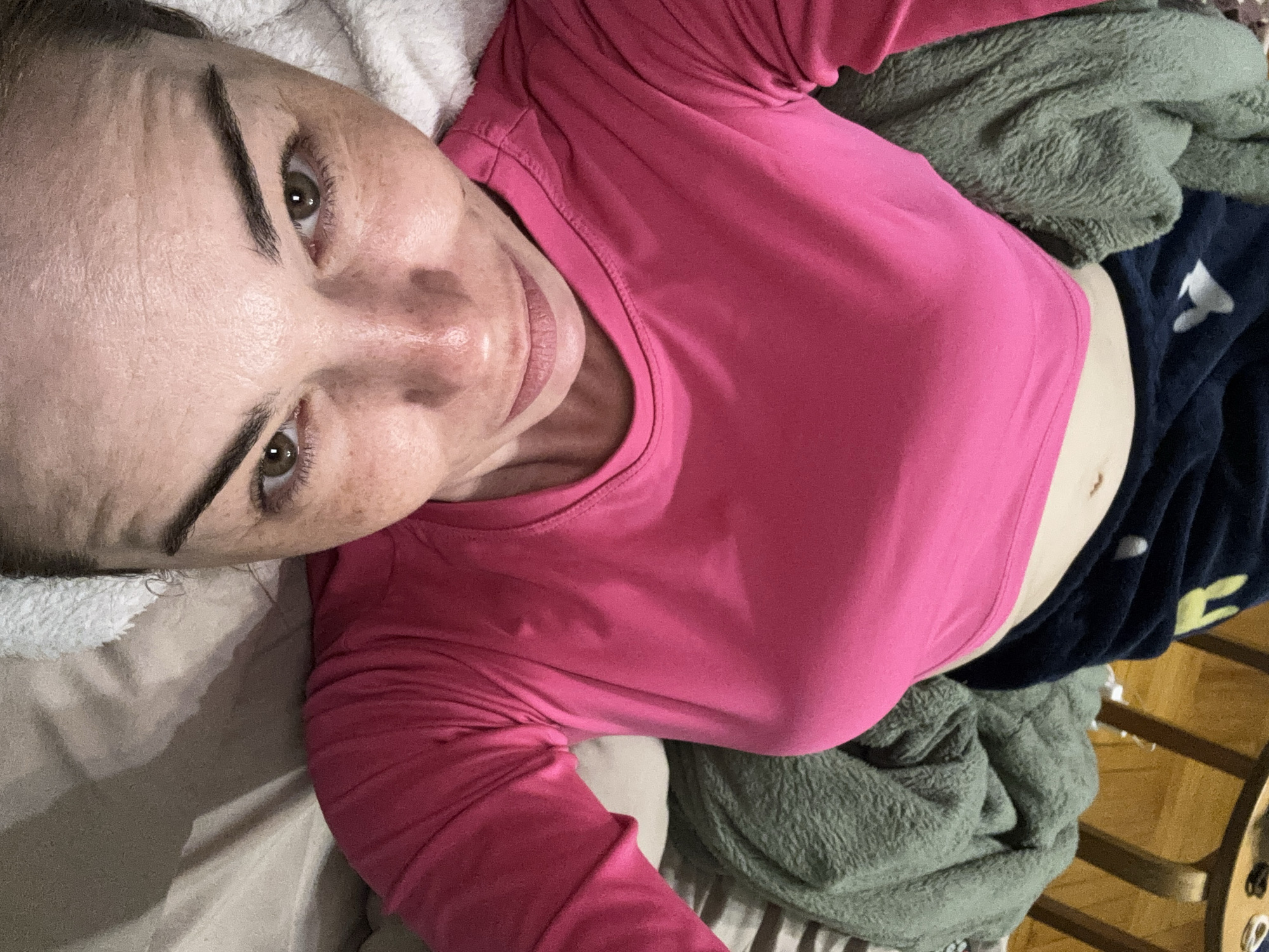 A fair-skinned woman with freckles is lying on her back, partially covered with a blanket and wearing a pink long-sleeved shirt and navy blue shorts with white graphics. Her head rests on a white towel or pillow. The background includes a green fleece blanket and the edge of a wooden table. Location: Unknown, Russia.