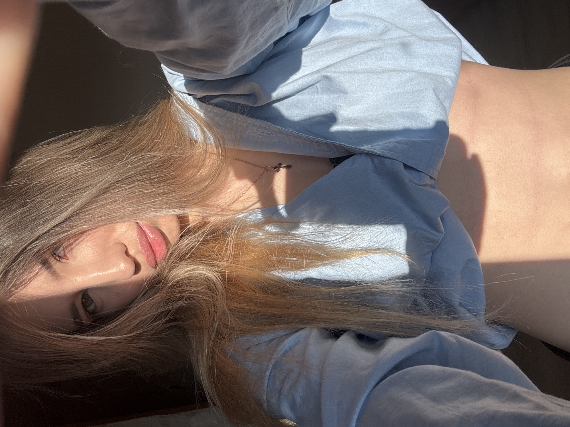 A woman with blonde hair is lying on her side, wearing a light blue shirt. A gold necklace with a cross pendant is visible on her chest. The scene is bathed in sunlight, creating strong shadows on her face and shirt. The location, Noviy, Russia, cannot be determined from the image.
