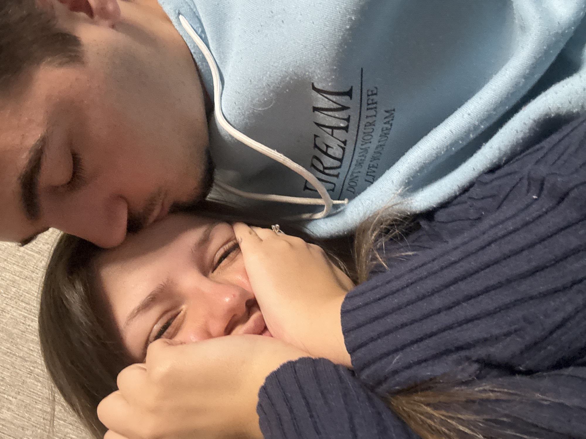 A man is kissing a woman on the forehead. The woman is smiling and has her hands near her face. The man is wearing a light blue hoodie with white drawstrings and a black inscription that reads "DREAM DON'T DREAM YOUR LIFE LIVE YOUR DREAM". The woman is wearing a dark blue sweater. The background is a light-colored surface. There is no indication of this image being taken in Sofia, Bulgaria.