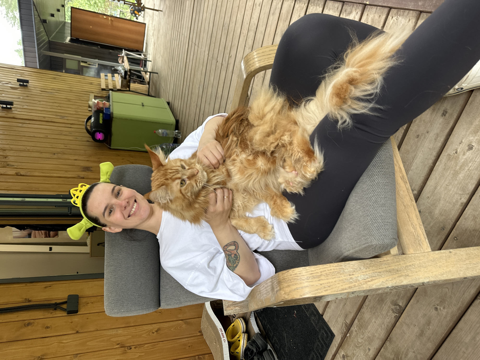 A woman with a Shrek-themed headband and a crown is sitting in a gray upholstered chair on a wooden deck. She is holding a large, fluffy orange cat on her lap. The woman is smiling and wearing a white t-shirt and black leggings. A tattoo is visible on her left forearm. To the left of the chair, there is a cat scratching post with yellow and black shoes on it, and a black rug. In the background, there is a wooden wall with doors, a green cabinet, and some outdoor furniture. The setting appears to be a residential outdoor space, possibly a porch or patio, in the city of Perm, Russia.