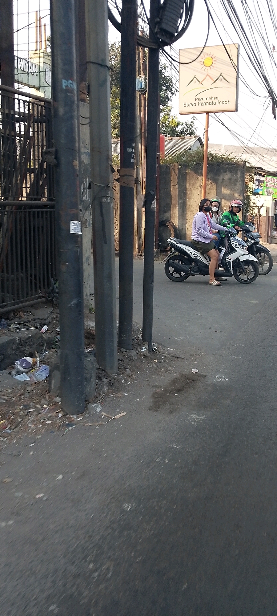 The image shows a street scene in Bekasi, Indonesia. In the foreground, several utility poles line the side of the road, with wires running overhead. Debris and trash are visible at the base of the poles. A white motorcycle is in motion on the asphalt road, carrying two passengers. The passenger in the front is wearing a light purple long-sleeved shirt and dark shorts, with a white mask and a helmet. The passenger behind her is wearing a green helmet and a green jacket. A second motorcycle, darker in color, is partially visible behind the first. In the background, a sign reads "Perumahan Surya Permata Indah," indicating a housing complex. Further back, a building with a tiled roof is visible. The sky is bright, suggesting daytime.