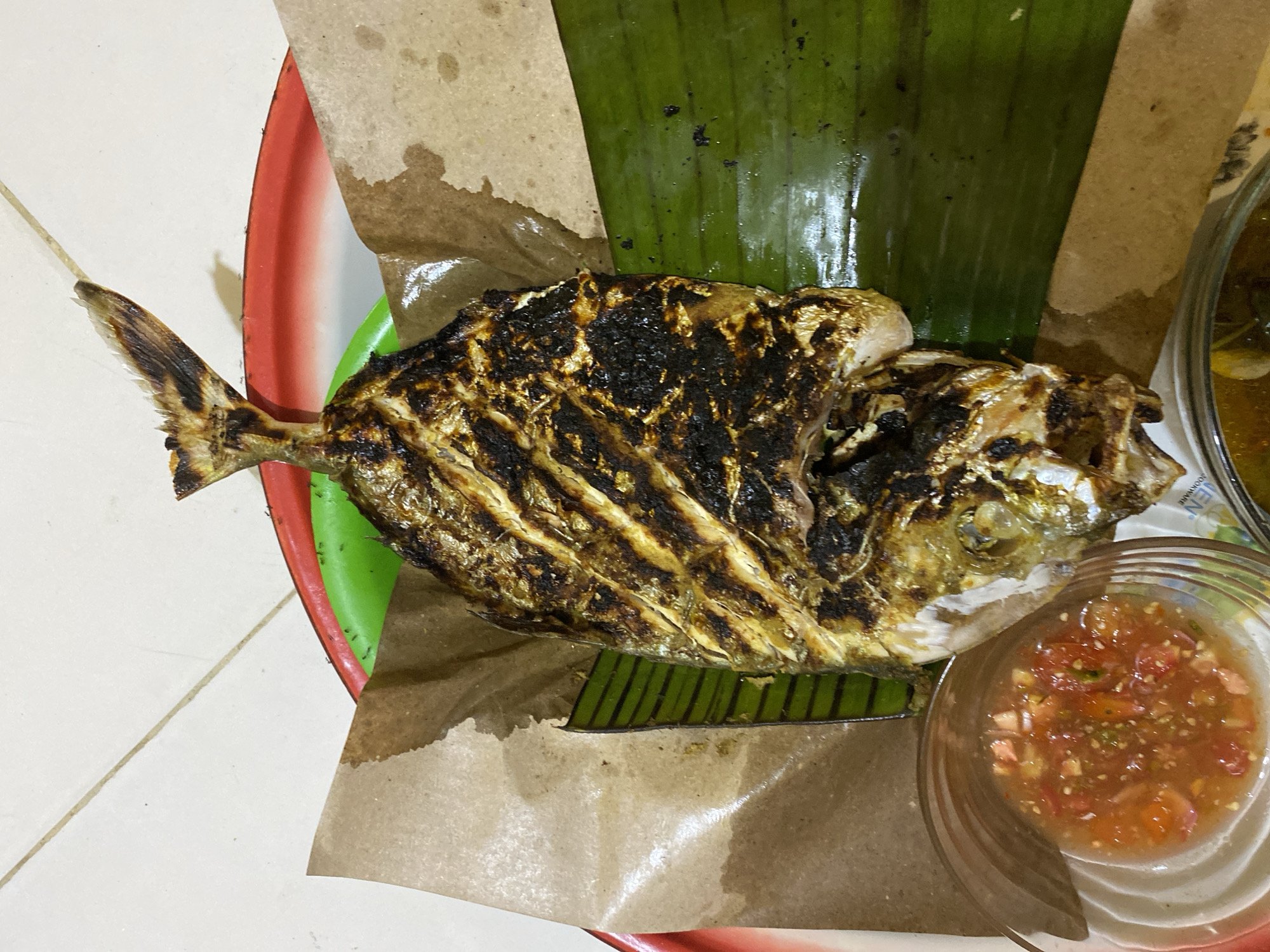 A high-angle, close-up photograph captures a whole grilled fish, likely a trevally, served as part of a meal. The fish, oriented horizontally with its tail pointing left, dominates the center of the frame. It displays distinct char marks across its skin, indicating it has been grilled, giving it a dark brown and golden-brown, somewhat glossy finish.
The fish rests on a bed composed of a large, dark green banana leaf and a piece of brown parchment paper. Both the leaf and paper are placed on a red and green plastic tray, which is largely obscured. The parchment paper shows signs of grease absorption, particularly around the fish.
To the lower right of the fish, a small, clear glass bowl contains a vibrant red salsa or sambal, featuring visible chunks of tomato, chili, and possibly onion in a liquid base. A portion of another bowl, holding a yellowish liquid that could be soup or another sauce, is visible in the upper right corner.
The setting appears to be a casual dining environment, possibly indoors or semi-outdoors, with the tray positioned on a light-colored tiled floor. Subtle details include a few small ants visible on the green part of the plastic plate near its red rim, and faint, partial, and illegible text on the rim of the glass bowl. The presentation style, using a banana leaf and a chili-based dipping sauce, is characteristic of Indonesian cuisine, aligning with the Tanjongkapai location context. No people are present, and the time of day is not discernible.