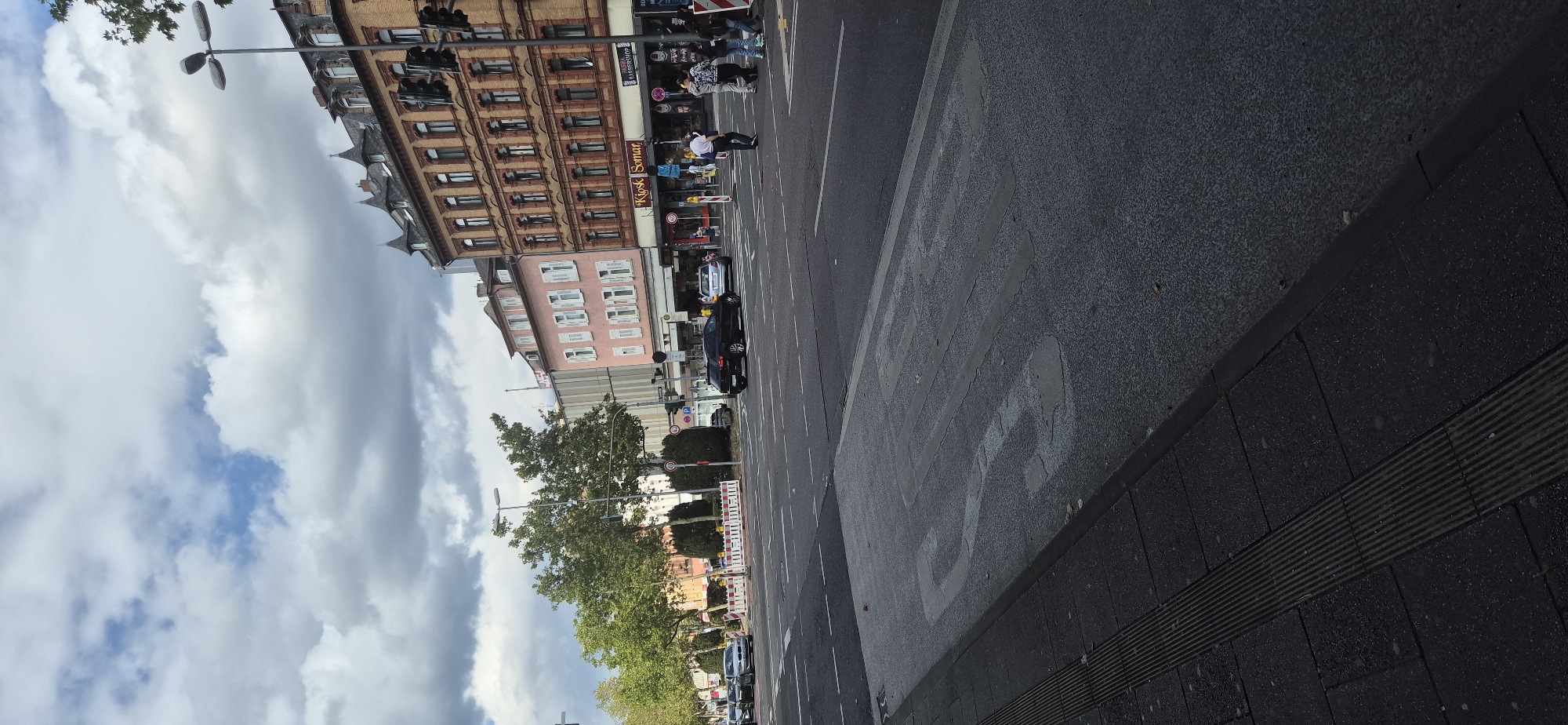 The image depicts a street scene in Wiesbaden, Germany. In the foreground, a speed bump with faded markings for "30" is visible on the asphalt road. A pedestrian sidewalk with tactile paving runs along the left edge of the road.
The street is a multi-lane intersection with traffic lights and road markings. Several cars are present, including a black SUV and a silver sedan driving on the road. Pedestrians are crossing the street at a designated crosswalk.
On the right side of the street, there are several buildings with commercial establishments. One building is pink and has multiple windows. Another building is made of brick and features a sign for "Kiosk Somar" and "Barbershop." Traffic lights are visible mounted on poles.
The sky is cloudy with patches of blue sky peeking through. Trees line the left side of the street, and traffic signs are visible. The overall scene appears to be a typical urban environment with ongoing traffic and pedestrian activity.