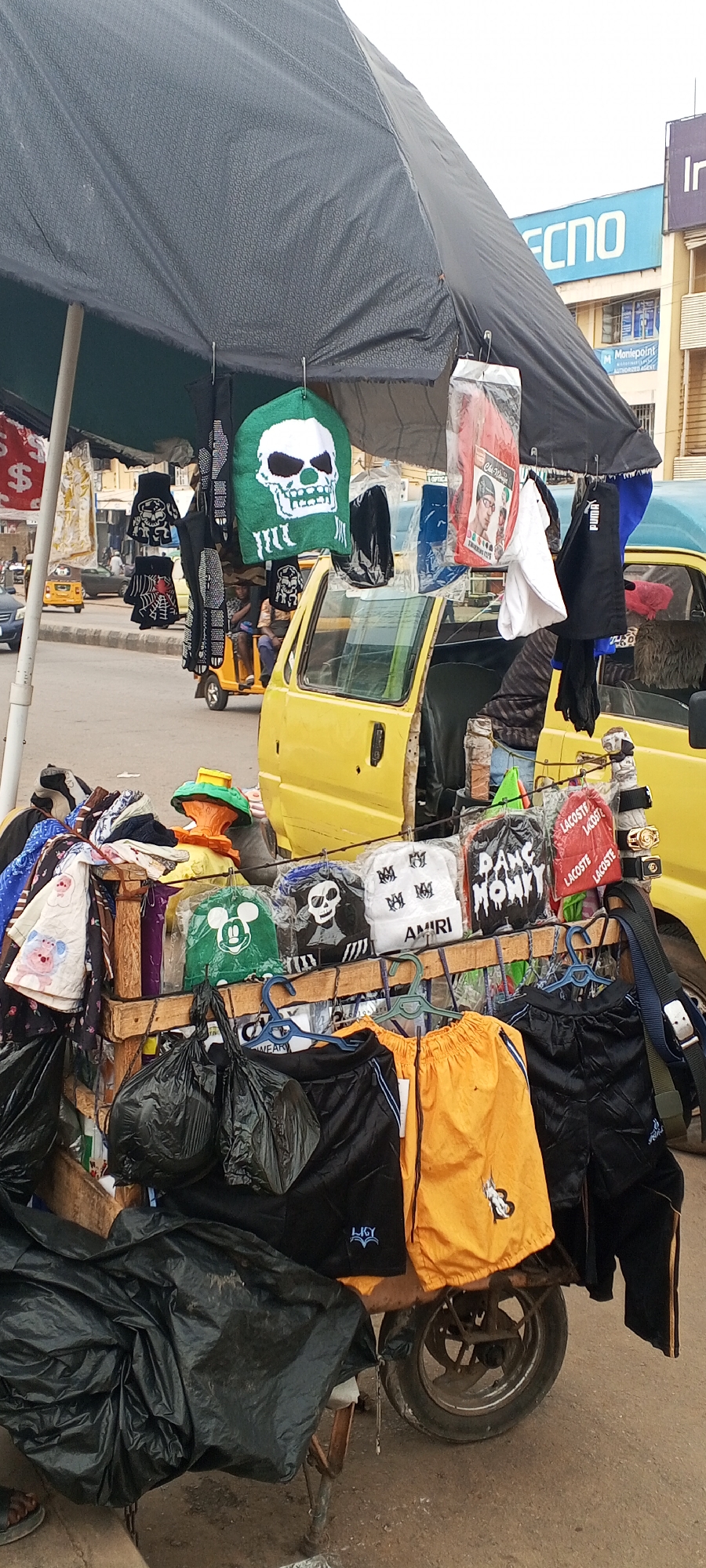The image depicts a street vending stall in what appears to be Maiburiji, Nigeria. The stall is set up on a wheeled cart, displaying various clothing items such as hats, scarves, and shorts, all hanging from hangers. A black umbrella provides shade to the items. The cart is positioned near a yellow vehicle, possibly a small van or tricycle. In the background, city streets are visible with other vehicles and a building with a "TECNO" sign. A secondary building with the words "Montepoint" and "Authorised Agent" is also visible. There are a few people visible in the background in a yellow tricycle. The ground appears to be paved.