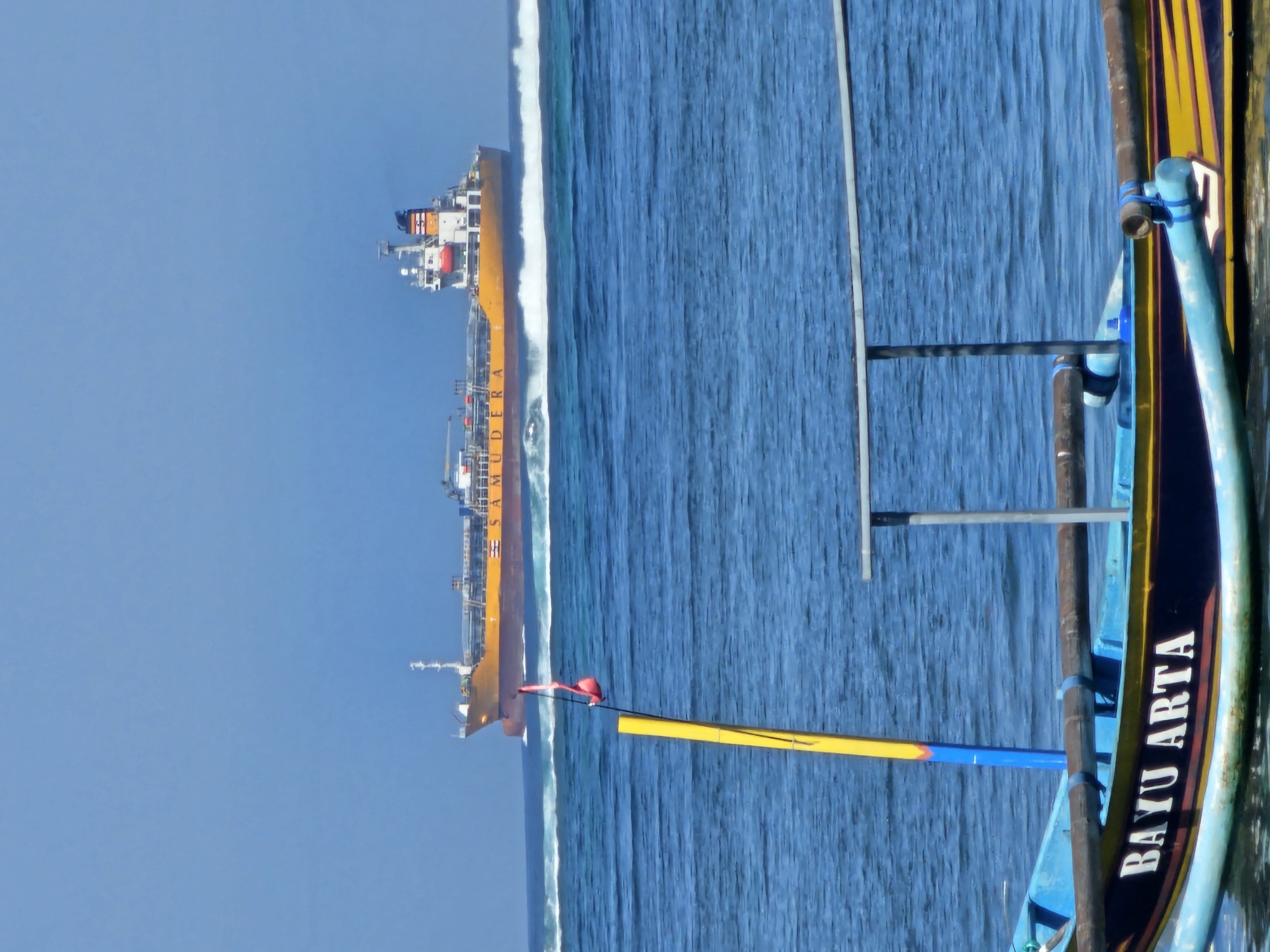 A bright, sunny day scene captures the ocean off Denpasar, Indonesia. In the foreground, the bow of a small, blue and yellow fishing boat named "BAYUARTA" is visible, with wooden outriggers and a yellow pole with a red flag. Behind it, a large orange tanker ship sails on the horizon, leaving a wake. The water is a vibrant blue, with small waves on the surface. The sky is a clear, cloudless blue. The composition emphasizes the vastness of the ocean and the contrast between the small fishing boat and the large commercial vessel.