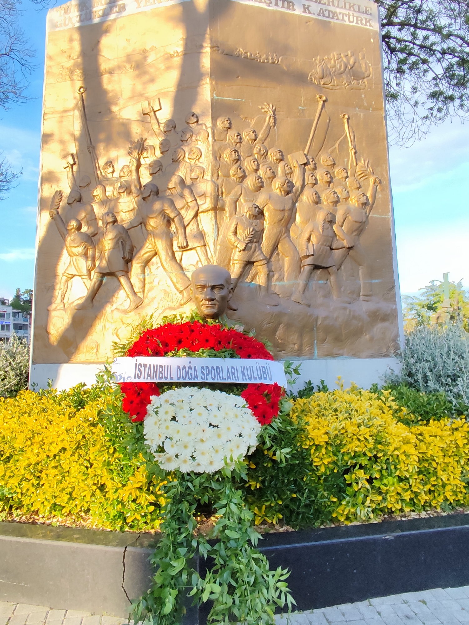 The image shows a monument dedicated to Mustafa Kemal Atatürk in an unknown city. The monument is a large bronze relief depicting scenes of Turkish history and revolution. At the base of the monument, a wreath of red and white flowers has been placed, with a banner that reads "İSTANBUL DOĞA SPORLARI KULÜBÜ". The monument is surrounded by colorful bushes and greenery. The environment appears to be a public park or memorial site.