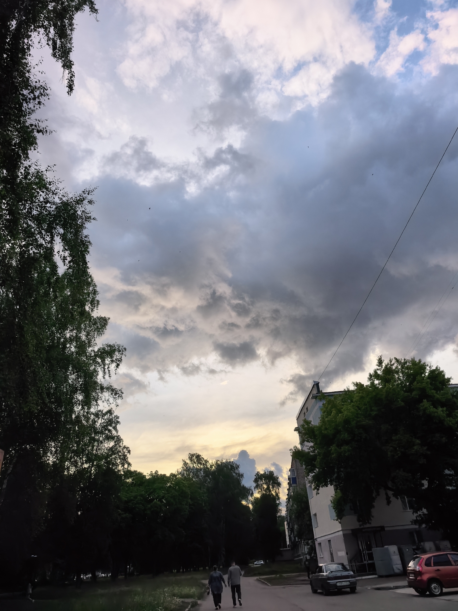 The image depicts a street scene in what appears to be a residential area in Sterlitamak, Russia. The sky is overcast with heavy, gray clouds, though a hint of lighter sky is visible near the horizon. Trees line the street on both sides, with lush green foliage obscuring the buildings behind them in some areas.
Two people are walking on the sidewalk, seemingly side by side. Several cars are parked along the side of the road, including a red hatchback. A tall building with an off-white facade is partially visible on the right side of the frame. An overhead electrical line cuts diagonally across the sky. The overall impression is one of a cloudy, possibly late-afternoon or evening scene.