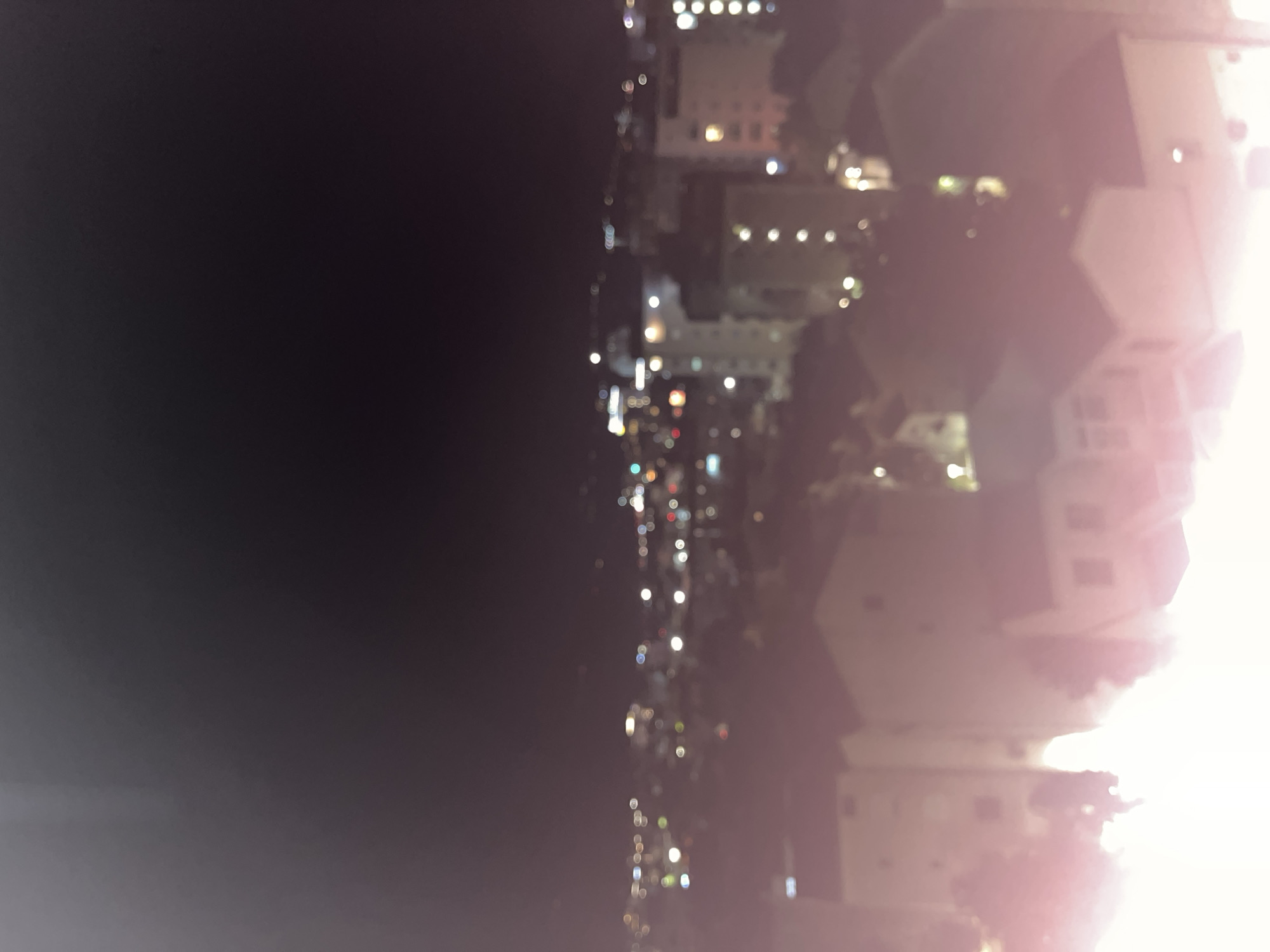 The image is a nighttime, out-of-focus photograph of a city skyline, likely Budva, Montenegro. The foreground is dominated by the bright, blown-out lights of what appear to be buildings and houses. In the mid-ground, a hazy collection of illuminated buildings with visible windows suggests a denser urban area. The background shows more distant, blurred lights, indicative of further development or infrastructure. The sky is dark and black, typical of nighttime. The overall impression is one of an urban landscape at night, with the blur obscuring specific details and creating a soft, ethereal glow.