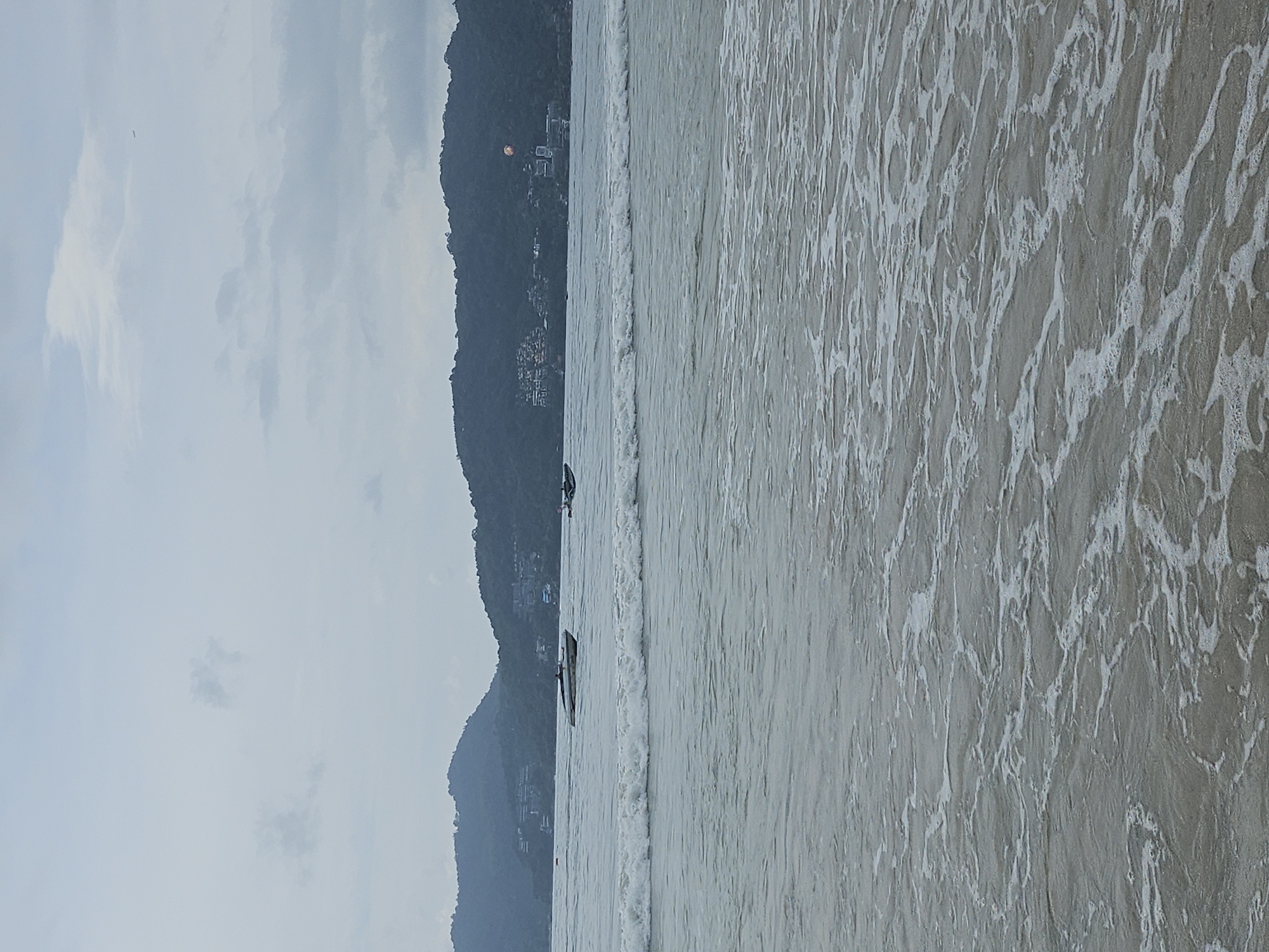 The image depicts a cloudy day at Patong Beach, Thailand. The foreground is dominated by the gentle waves of the ocean washing onto the sandy shore, with white foam creating intricate patterns on the water's surface. In the mid-ground, several boats are visible on the sea. One speed boat is closer to the viewer, and further out, another smaller boat is present. In the background, a lush, green, mountainous coastline lines the horizon. Buildings, likely hotels or resorts, are nestled into the hillsides, indicating the populated nature of the area. A parasail, with an orange and yellow canopy, is visible against the backdrop of the mountain, suggesting recreational activities. The sky is overcast with diffused light, creating a muted color palette across the entire scene.