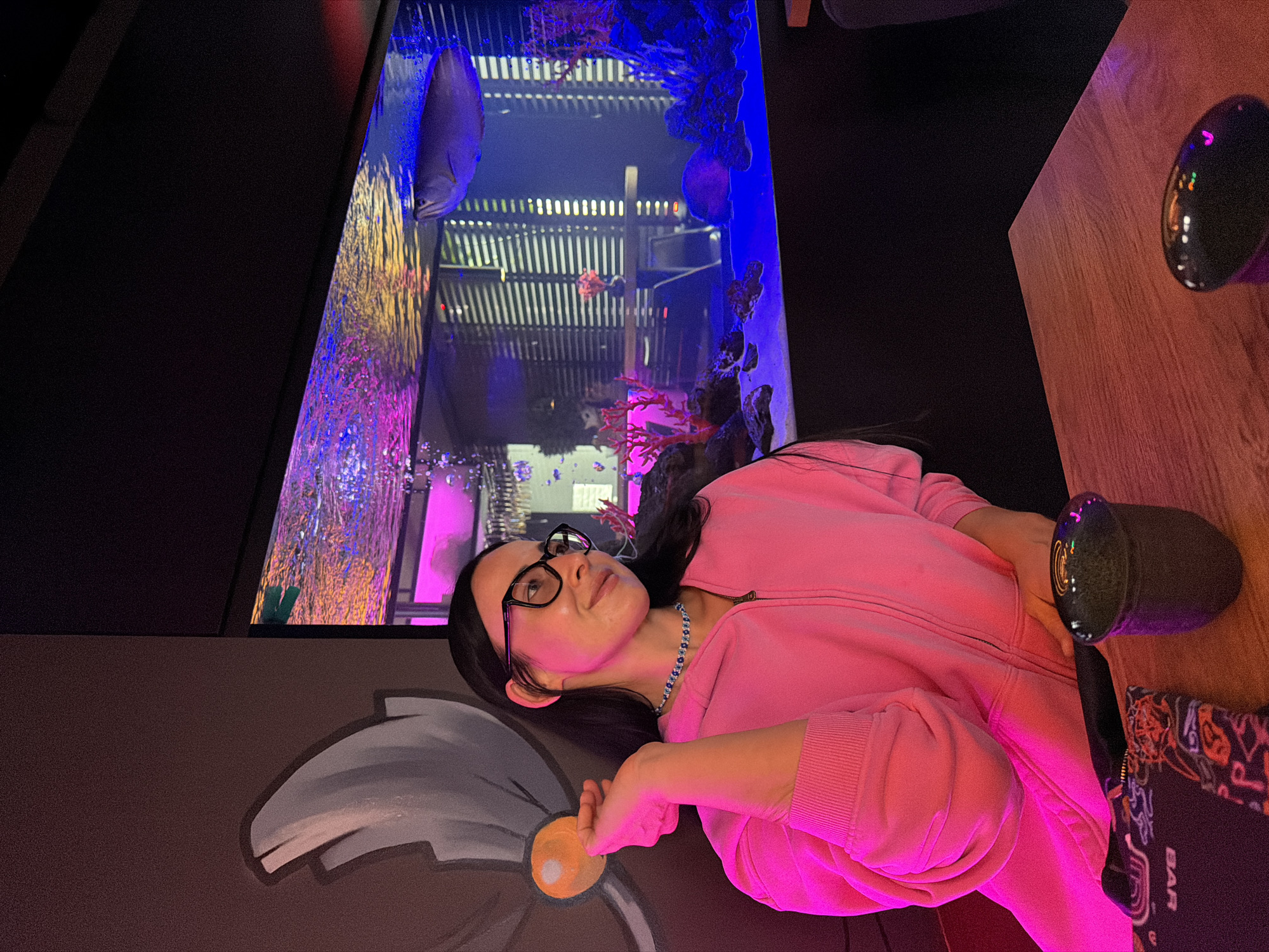 An indoor scene in Saint Petersburg, Russia, depicts a woman enjoying the ambiance of what appears to be a modern restaurant or bar. The image is rotated 90 degrees clockwise. The central figure is a woman with dark hair, wearing black-rimmed glasses, a bright pink hooded zippered sweatshirt, and a light blue and white beaded necklace. She looks upward and slightly to her right with a gentle expression, possibly a faint smile, her left hand resting near an orange circular element on a dark mural.
The dominant feature of the setting is a tall, narrow aquarium illuminated with vibrant artificial lighting in shades of blue, purple, and yellow. Large and small fish, including a prominent greyish fish near the top left, swim amidst coral and other aquatic decorations. Reflections on the aquarium glass show a slatted ceiling or background structure. The overall lighting is dim, suggesting an evening or nighttime setting.
To the right, a wooden tabletop is visible, holding a dark, possibly metallic, cup. Next to the cup, a dark item, likely a menu or coaster, displays colorful patterns and the word "BAR" in white text, rotated. The wall behind the woman features a large, abstract mural in grey and white tones, complementing the modern decor. The woman appears relaxed, observing the aquatic display within the illuminated tank.