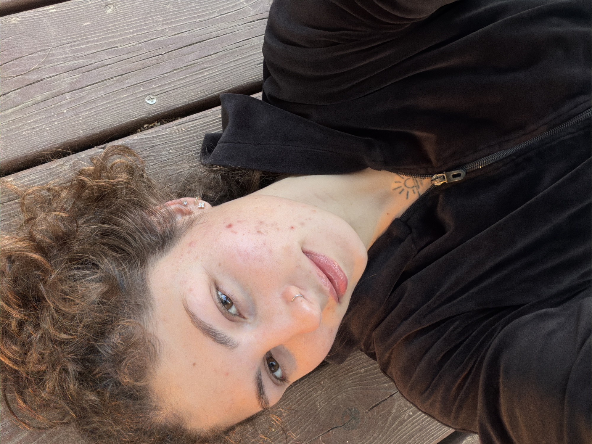 A young person with curly brown hair is lying on their back on a weathered wooden surface. They are wearing a black zipped-up velvet jacket. Their skin is fair, with some visible acne. They have brown eyes and a nose ring. A small tattoo of a sun is visible on their neck. The background is blurred, suggesting a shallow depth of field. The location is not evident from the image, therefore I cannot determine if the image was taken in Kiryat Bialik, Israel.