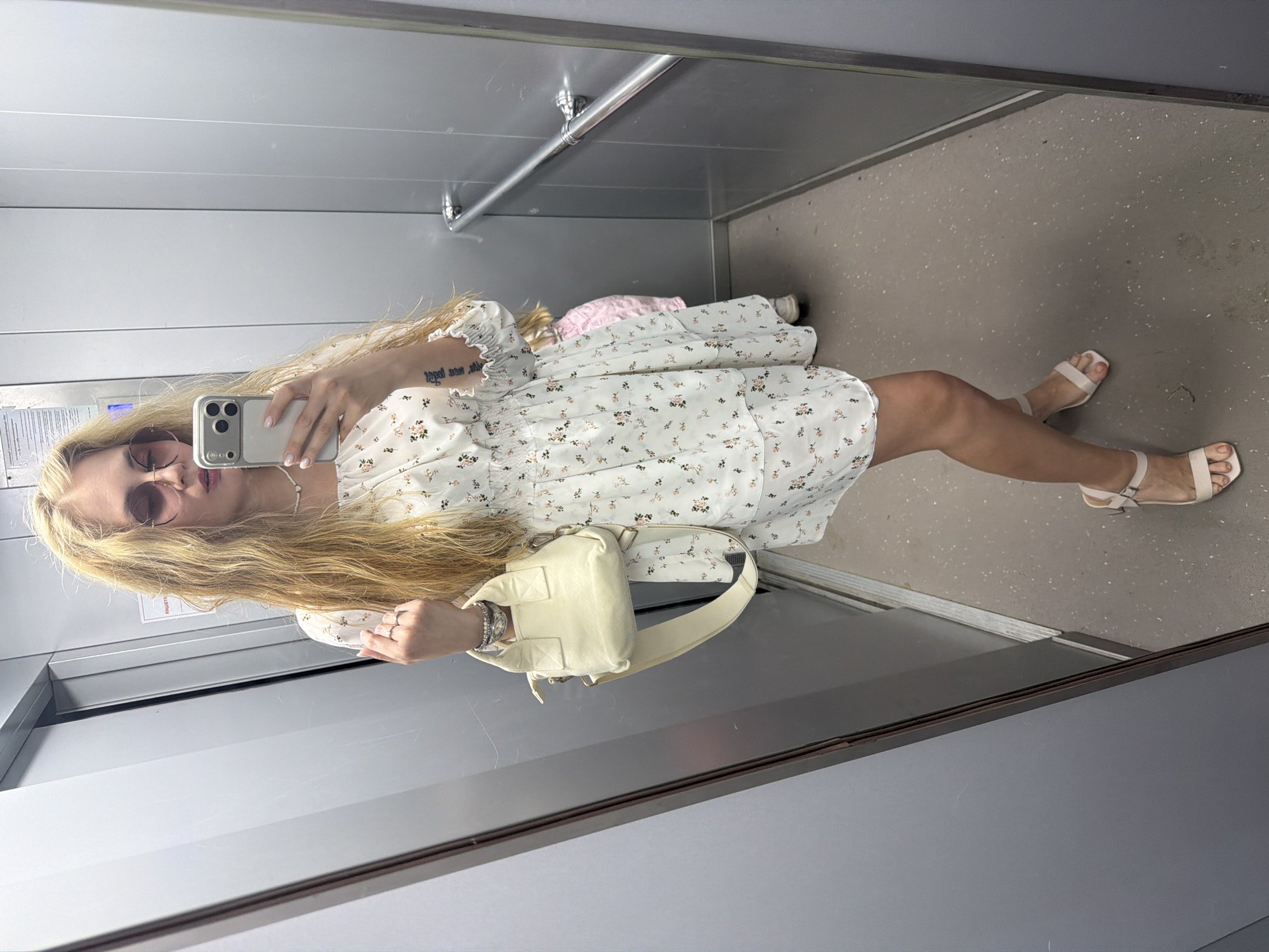 A blonde woman with sunglasses is taking a selfie in an elevator. She is wearing a short, white floral dress and light pink sandals. She carries a small, cream-colored purse. The elevator has metal walls and a gray floor. A handrail is visible along the upper wall. The location is unknown, and there is no discernible evidence to suggest the city of Chaikovsky, Russia.