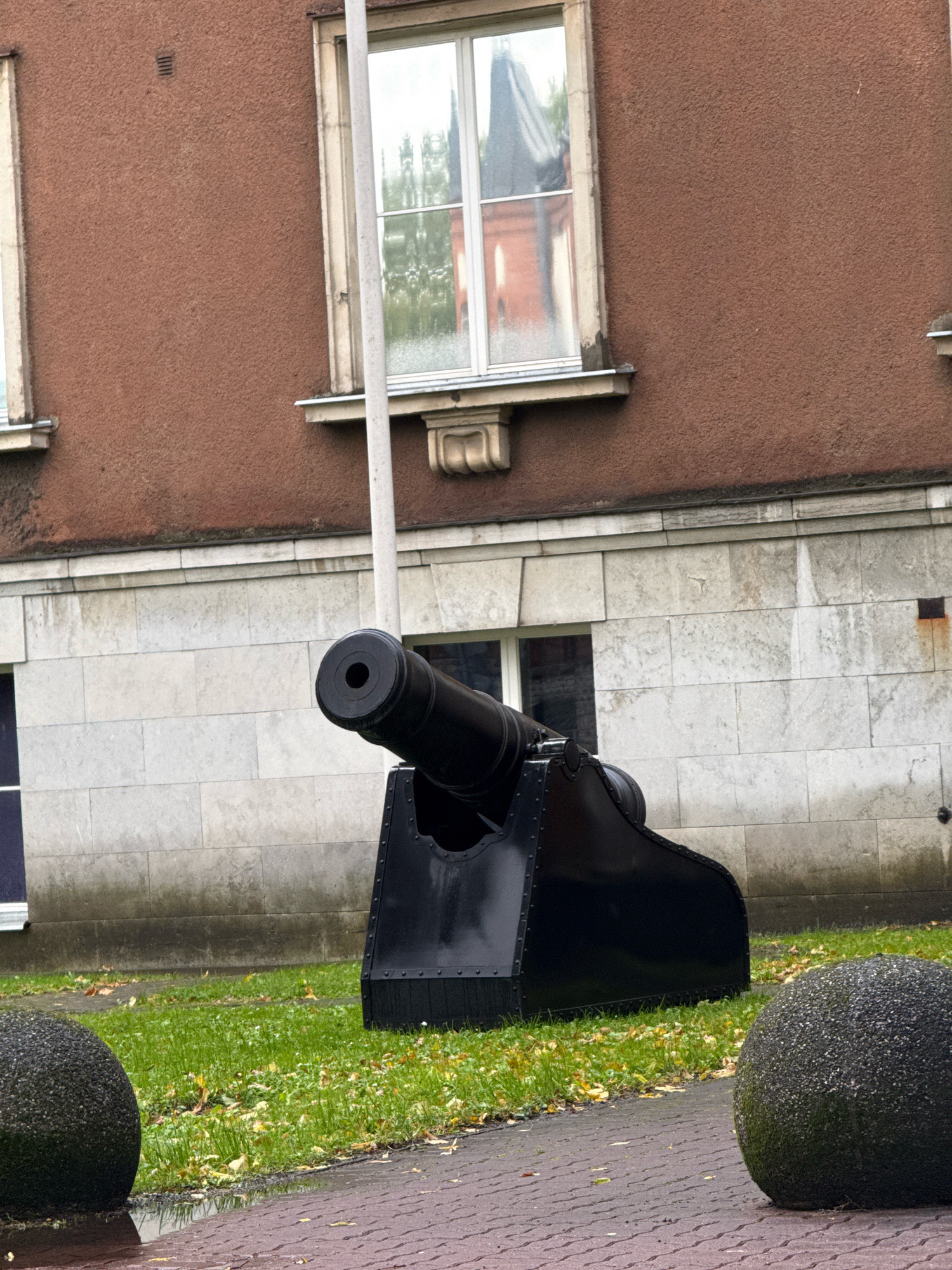 The image depicts an outdoor scene in Tallinn, Estonia, featuring a historical cannon positioned in front of a building with a textured brown upper facade and a lighter-colored stone lower facade. The cannon is black and rests on a dark carriage with visible rivets. In the foreground, there is a paved walkway with a reddish-brown brick pattern, a patch of green grass with fallen autumn leaves, and two large, dark, spherical bollards. The building has large windows with reflective glass, showing reflections of trees and architecture. A tall, thin, white pole is situated vertically to the right of the cannon. The overall atmosphere suggests a historical or commemorative setting.