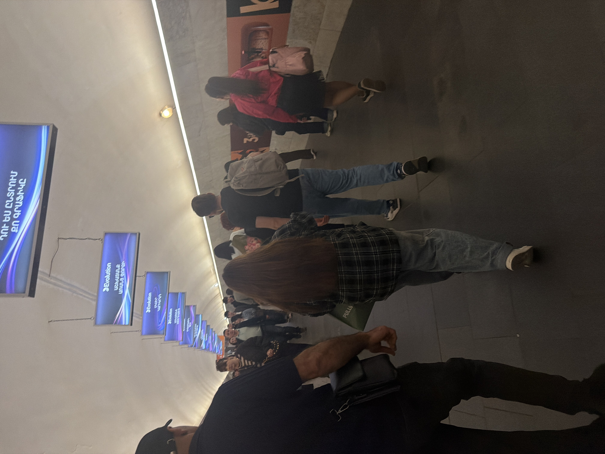 This image shows a crowded corridor in what appears to be a subway station in Yerevan, Armenia. People are walking in the same direction. Several small, rectangular electronic signs hang from the ceiling on the left, and the largest sign on the left displays text in Armenian, reading "ԴՈՒ ԵՍ ԸՆՏՐՈՒՄ ՔՈ ԳՐԱՖԻԿԸ" which can be translated to "You choose your schedule". Another sign contains the text "Evolution" and "ԱՇԽԱՏԱՆՔ ԱՌԱՆՑ ՓՈՐՁԻ" translated to "work without experience".
A person with a black cap, black t-shirt, and dark pants is in the lower right foreground of the image.