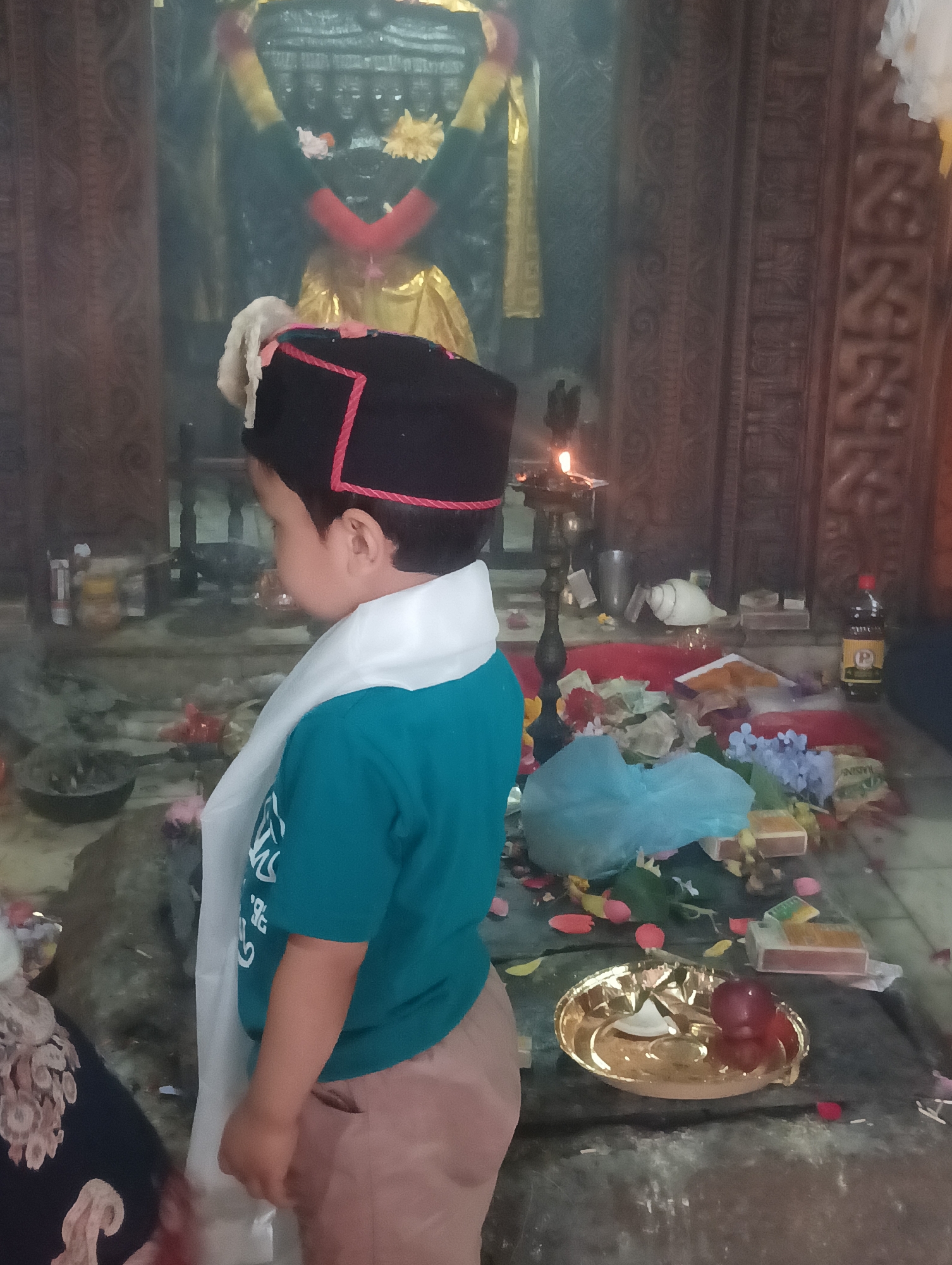 A young boy, facing away from the camera, stands in what appears to be a temple or shrine. He is wearing a black cap with red trim and a white scarf draped over his shoulders, over a teal t-shirt and tan pants. The boy is looking towards an altar area that is adorned with offerings.
The altar is cluttered with various items, including flowers (some real, some artificial), small boxes, a conch shell, and what looks like a bottle of dark liquid with a yellow label. A small oil lamp with a flame is visible, suggesting a religious ceremony or ritual is taking place, likely in the evening or a dimly lit interior. Petals are scattered on the floor around the altar.
Behind the altar, a dark, imposing structure with multiple carved faces or figures can be seen, likely a deity statue or religious iconography. The background also features wooden paneling with intricate carvings. The overall atmosphere seems reverent, with the focus on the boy and his interaction with the sacred space and its offerings.
The scene is set in Gojra, India. There is no visible text in the image that can be read clearly. The lighting suggests it is either dusk or an indoor setting with artificial light sources.