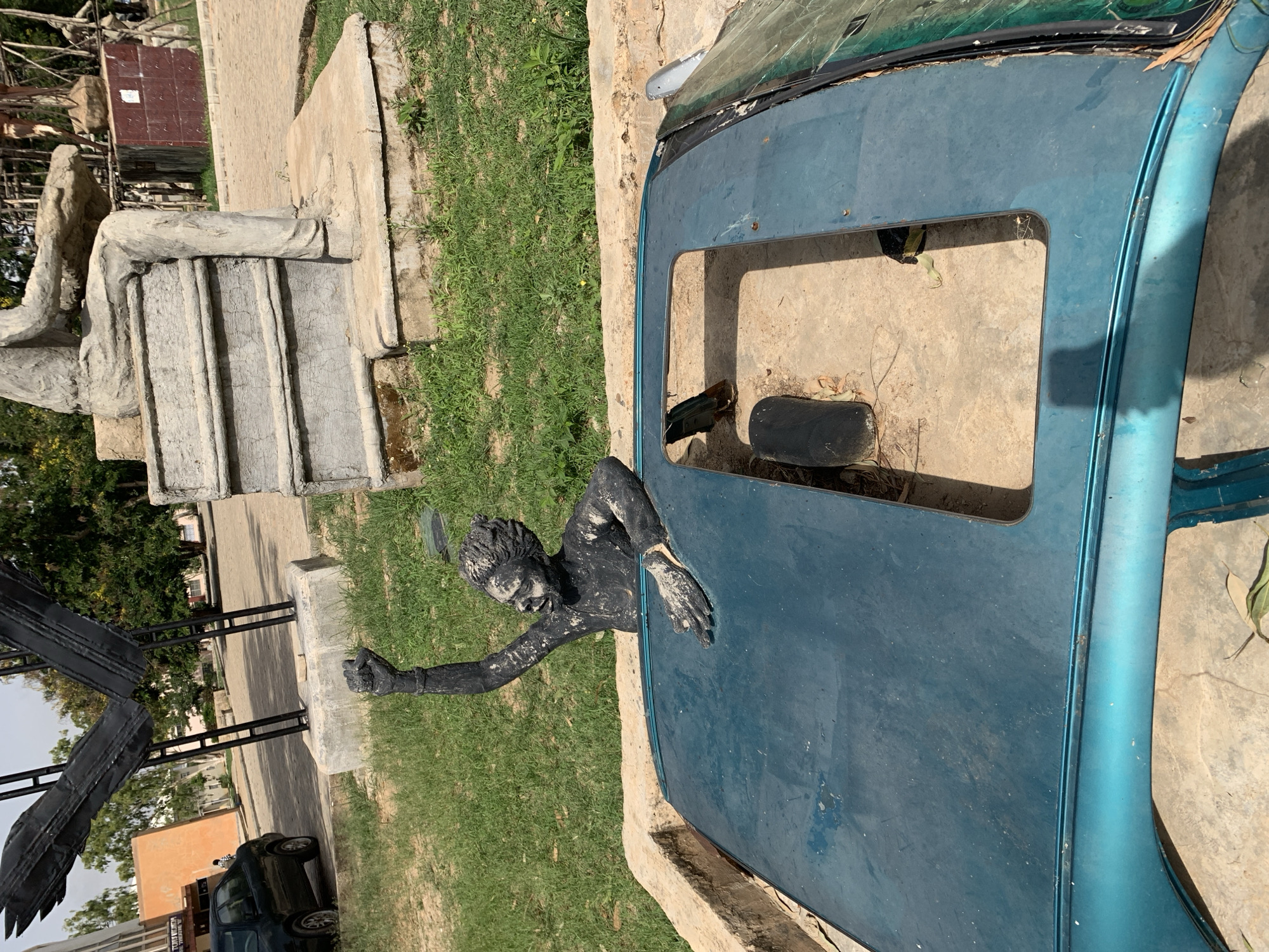 A public artwork stands in the city of Jos, Nigeria. The art piece features a dark-painted sculpture of a person emerging from an old, blue car roof with a sunroof, as if struggling to break free. The car roof lies on a concrete base surrounded by grass. In the background, other art installations, greenery, and a building are visible. A concrete monument resembling seated figure on stairs is partially visible in the background. The sky is overcast.