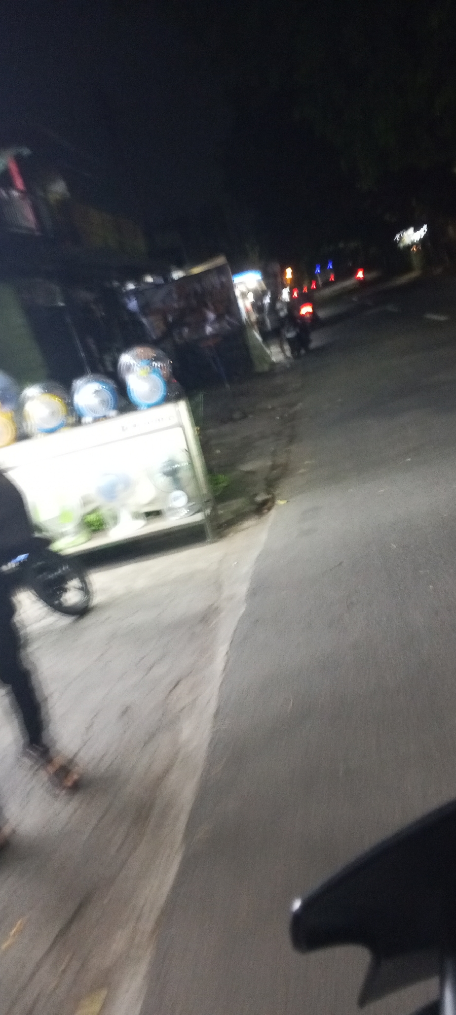 This is a blurry, night-time image taken from a moving vehicle, likely a motorcycle, on a street in Depok, Indonesia. The scene depicts a roadside stall selling various items, including what appear to be new electric fans displayed on a counter. To the left of the stall, a person's legs and feet are visible, wearing dark pants and sandals. A motorcycle is parked or moving alongside them. The stall itself is illuminated, casting light onto the surrounding pavement. Further down the street, several vehicles, including motorcycles, are visible, some with illuminated tail lights. The general environment appears to be a commercial or residential street, typical of urban areas in Indonesia at night. The focus is poor due to motion blur.
