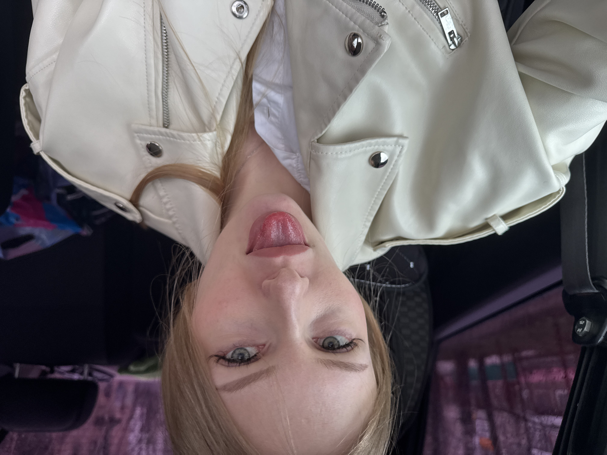 A fair-skinned young woman is posing for a picture, sticking out her tongue. She has blonde hair, green eyes, and defined eyebrows. She wears a white leather jacket over a white collared shirt. The scene appears to be inside a vehicle. Through the car window, a blurred cityscape is visible. Based on the location requested, the scene may be in Perm, Russia. However, there are no specific landmarks in the photo to confirm this.