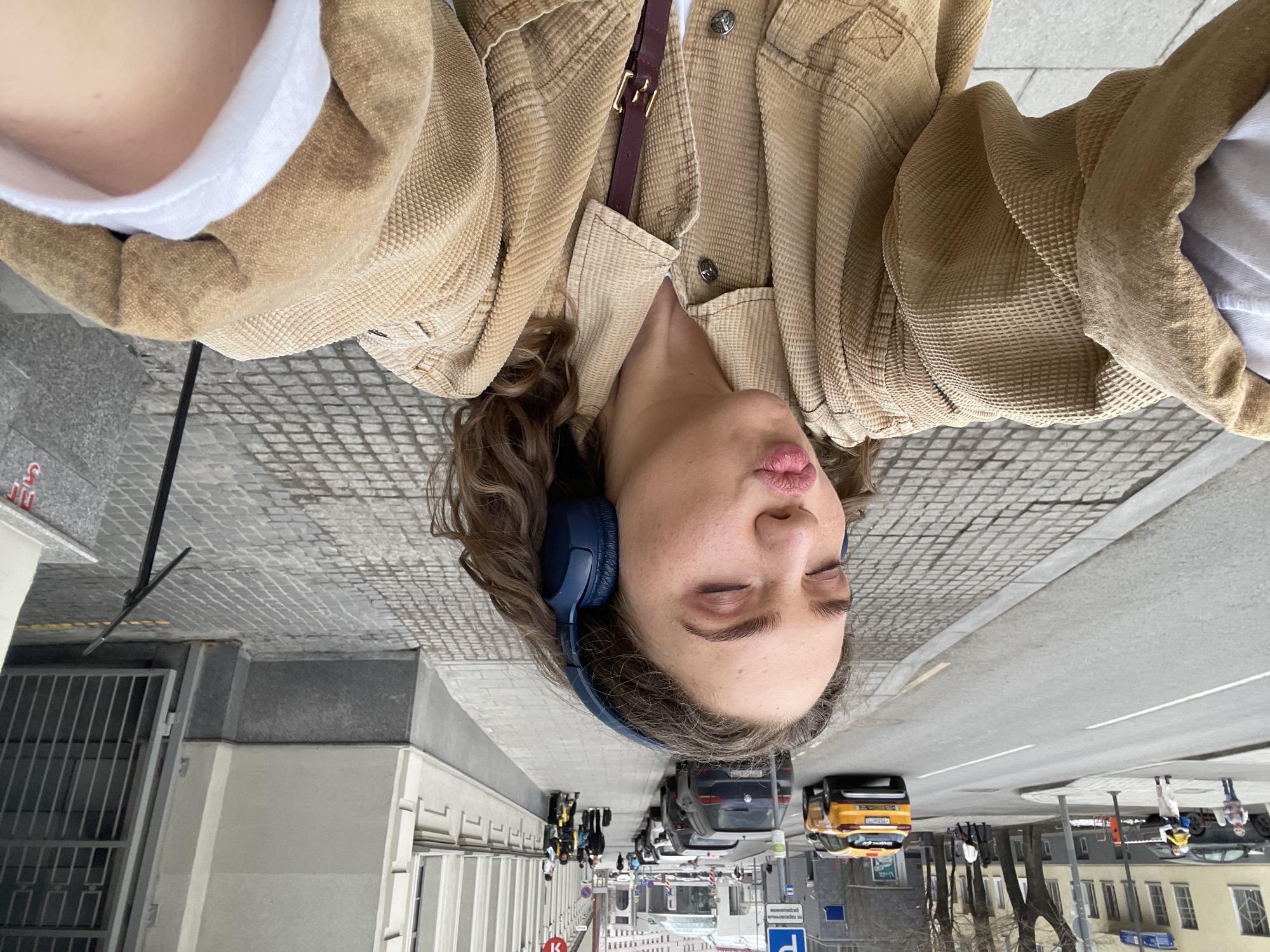 A selfie taken from an unusual perspective shows a young person wearing a light brown corduroy jacket, a white t-shirt, and blue headphones. The person has curly hair and is puckering their lips with their eyes closed.
The background reveals a street scene. Vehicles, including a yellow taxi and a dark-colored car, are visible. There are pedestrians on the sidewalk. The architecture includes buildings with light-colored facades.
Based on visual clues like signage written in Cyrillic and architectural styles, the location is Moscow, Russia.