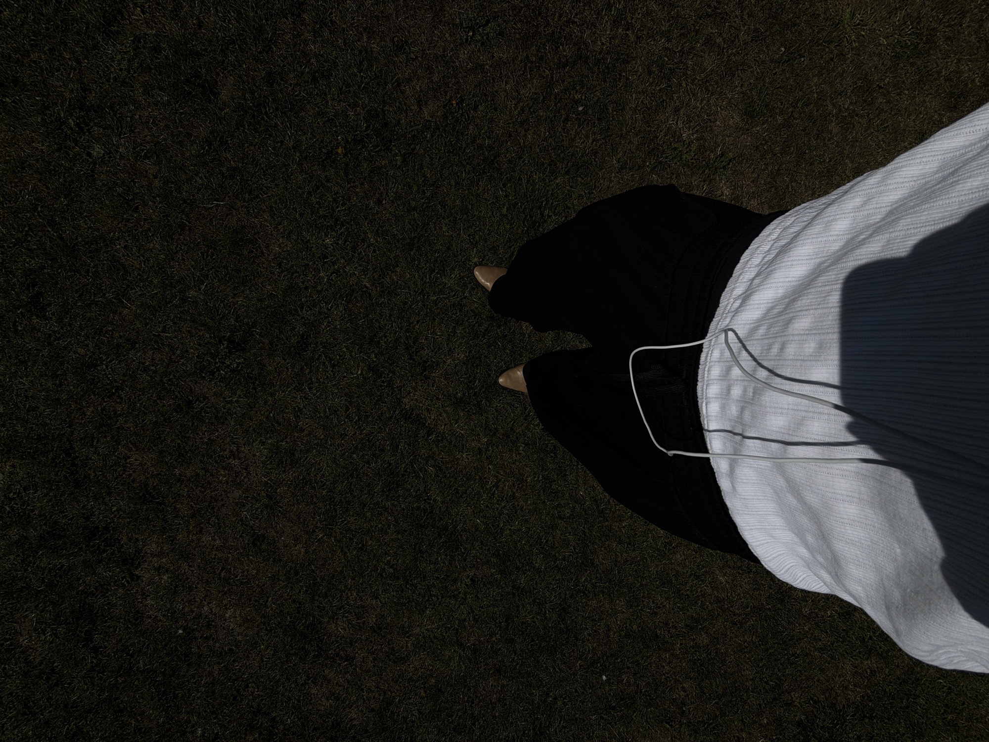 A top-down shot depicts a person standing on a lawn. The person is wearing black pants, beige shoes, and a white top. A white cord is visible near the person's waist. The background is a dark green lawn. The location is not identifiable as London, United Kingdom from this image.