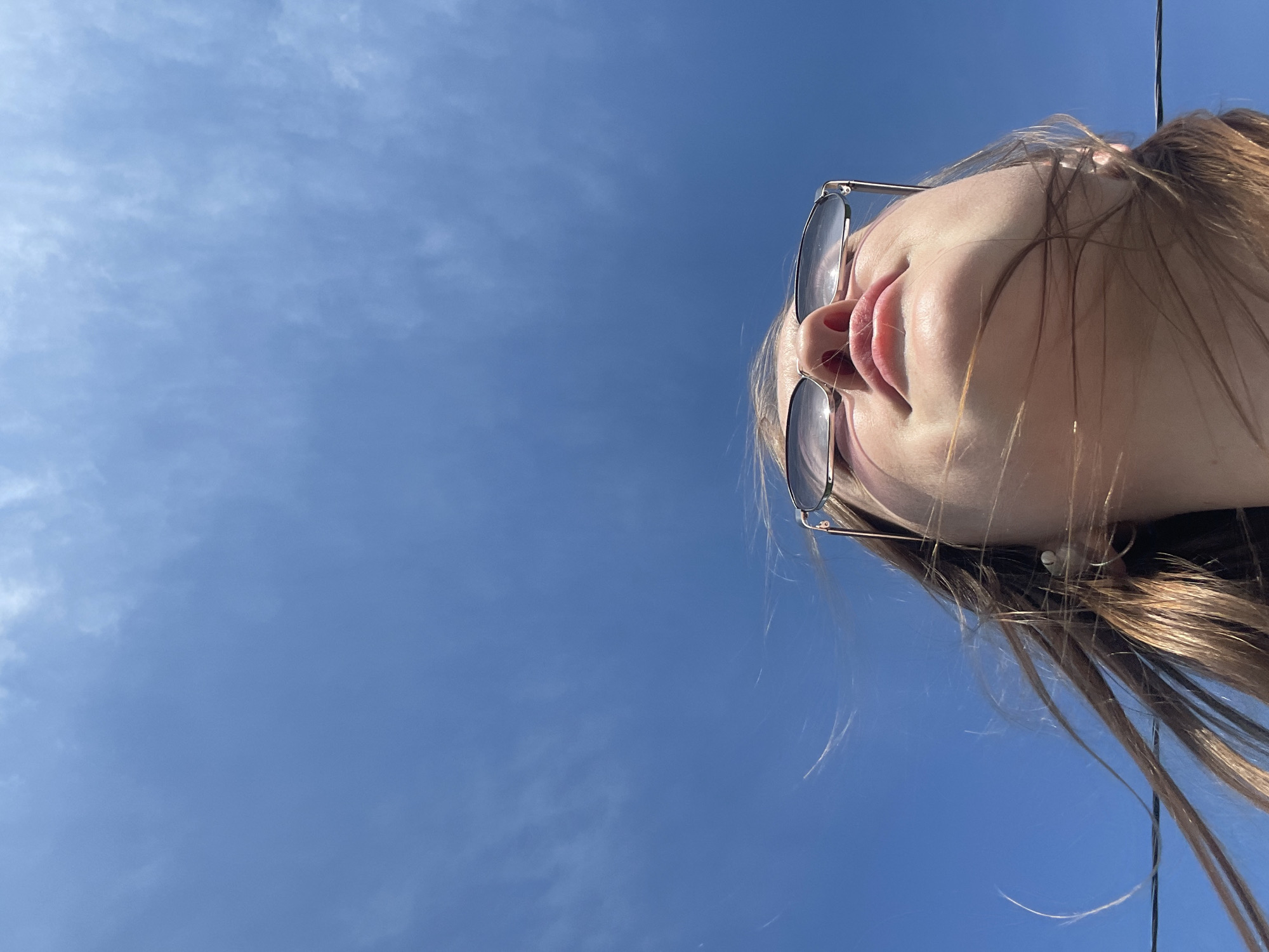 A low-angle, upward-looking portrait captures an individual, likely female, from the nose down to the upper chest, against a clear blue sky. The individual has light skin and visible lips, chin, and neck. Sunglasses with dark lenses and a thin metal frame are positioned on the bridge of the nose. Brown hair, possibly with lighter highlights, is visible and appears to be swept upwards and outward by wind, particularly around the jawline and below the chin. The dominant background is a bright blue sky, occupying approximately the upper two-thirds of the frame, with scattered, faint white cirrus-like clouds, especially noticeable in the upper right quadrant. A thin, dark, horizontal line, resembling a utility wire or cable, runs across the lower left portion of the frame, partially obscured by the individual's hair. The bright lighting indicates a sunny daytime setting. The image itself contains no discernible landmarks, architectural features, or geographical identifiers to visually confirm the provided location of Сырково, Russia. The scene depicts an open outdoor environment.
