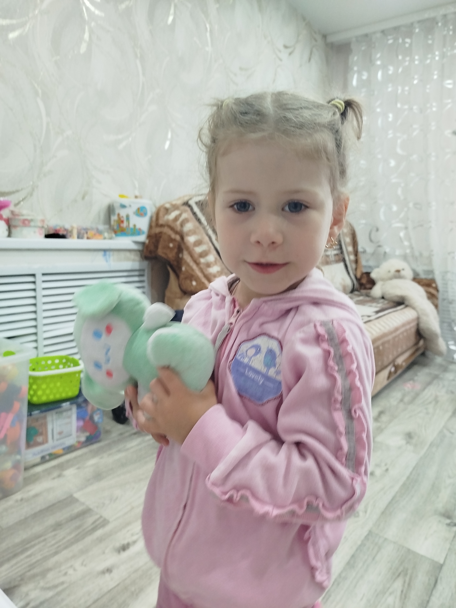 A young Caucasian girl with blonde hair pulled into two ponytails, stands in a room holding a green plush toy. She is wearing a pink hooded zip-up jacket with ruffled edges and a patch on the left chest. The room appears to be a living room or play area, with a couch in the background, a radiator, and shelves filled with toys. The floor is wooden. The image was taken in Krasny Gulyai, Russia.