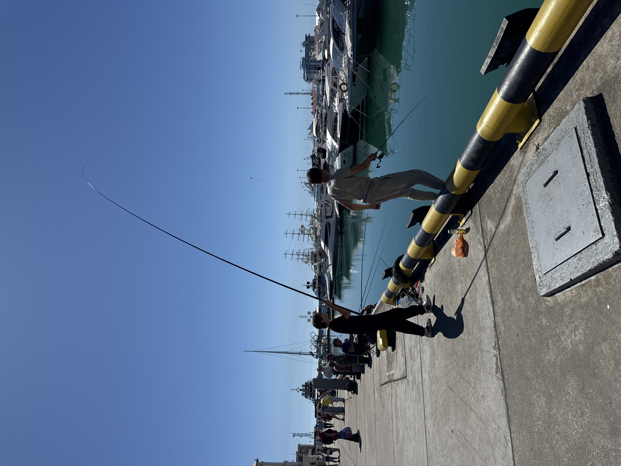 The image captures a lively, sunny day at a marina or pier in Sochi, Russia. The bright blue sky is clear, and the sun casts distinct shadows, indicating it is likely midday or early afternoon. The turquoise waters of the harbor are calm, reflecting the numerous luxury yachts and boats, including a large sailboat with multiple masts, docked along the pier.
In the foreground, two individuals are actively fishing. One person, dressed in light-colored attire, is balancing on a black and yellow striped safety barrier or bollard, holding a fishing rod out over the water. An orange plastic bag is hanging from this same barrier. To their left, another person, silhouetted in dark clothing, stands firmly on the concrete pier, also holding a fishing rod with its line cast into the water. Their shadow is visible on the textured concrete surface of the pier.
Further down the pier in the background, several other people are visible, many seated on portable chairs or standing, seemingly also engaged in fishing or observing the activity. A square metal hatch is visible on the concrete pier near the foreground. The scene depicts a relaxed, recreational atmosphere typical of a bustling harbor on a clear day. No discernible text is visible on any objects in the image.