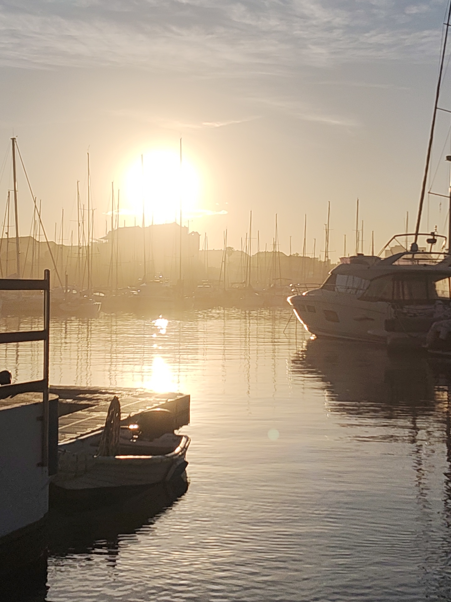 This image depicts a harbor scene in Dénia, Spain, at sunrise or sunset. The sun is positioned high in the sky, casting a golden glow over the water and the marina. In the foreground, a small boat is docked. The main subject is a modern yacht to the right, with smaller sailboats visible in the background, their masts creating a dense silhouette. The water is relatively calm, reflecting the sunlight and the shapes of the boats. In the background, a building, possibly a castle or fortified structure, can be seen on elevated ground. The overall atmosphere is serene and peaceful.