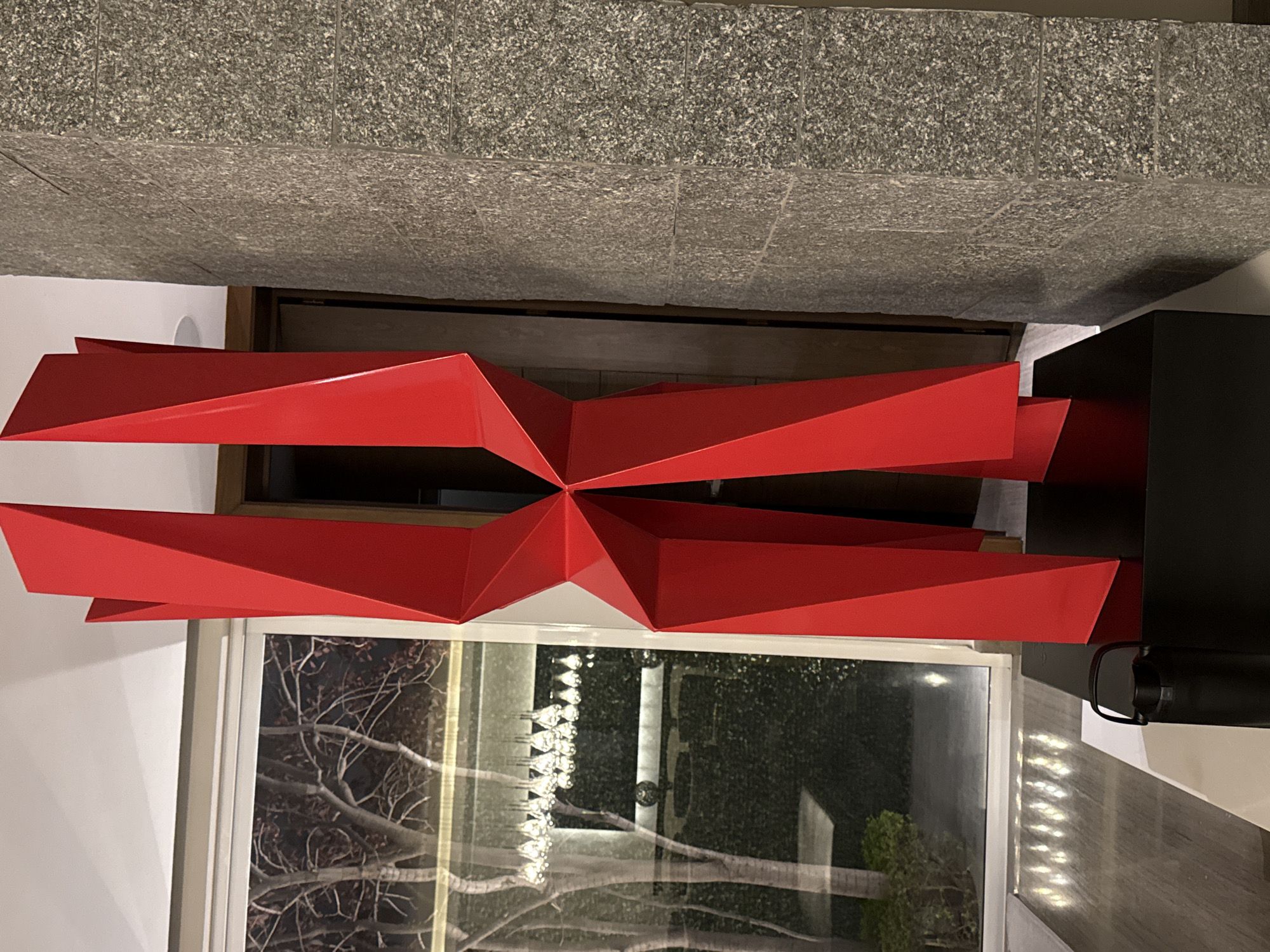 Captured indoors, the image presents a striking red, geometric art piece that seemingly floats above a window. The artwork, composed of angular segments converging at a central point, dominates the space. Below it, the window offers a glimpse of a tree with bare branches, possibly indicating winter or a late autumn scene, and a dark, possibly green background suggesting foliage or a wall. A strand of small lights is visible on the other side of the window. The top of the frame is dominated by a grey, granular surface, perhaps a wall or architectural element, while to the right, a black object protrudes into the scene. The setting appears to be a contemporary interior.