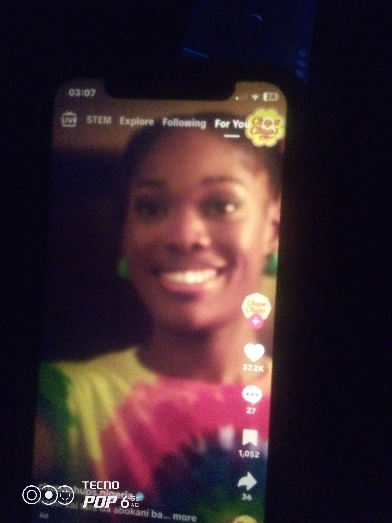 The image shows a close-up of a smartphone screen displaying a social media app, possibly TikTok. The app interface includes elements like "LIVE," "STEM," "Explore," and "Following," along with a "For You" section indicated by a Chupa Chups logo. A young Black woman is prominently featured on the screen, smiling. On the right side of the screen, there are social media interaction icons and counters, including a heart with "37.2K," a speech bubble with "27," a bookmark with "1,052," and an arrow with "36." The bottom of the screen shows a watermark or branding for "TECNO POP 6 4G." The background of the image is dark, suggesting the photo was taken in low-light conditions. There is no visible indication of the city of Howayi, Nigeria within the image itself.