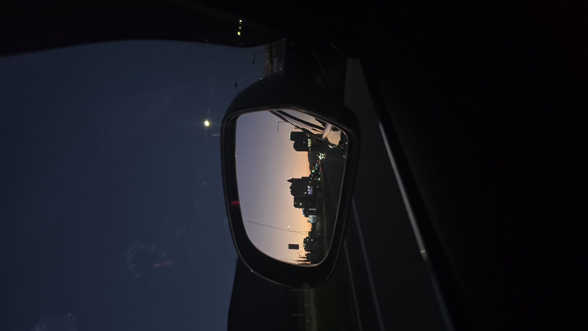 The image depicts a side view mirror reflecting a cityscape at dusk. The buildings are silhouetted against an orange and purple sky. Streetlights illuminate the road leading toward the buildings. The mirror is attached to a car. The scene is likely a highway or main road leading into the city. It’s not possible to identify the exact location.