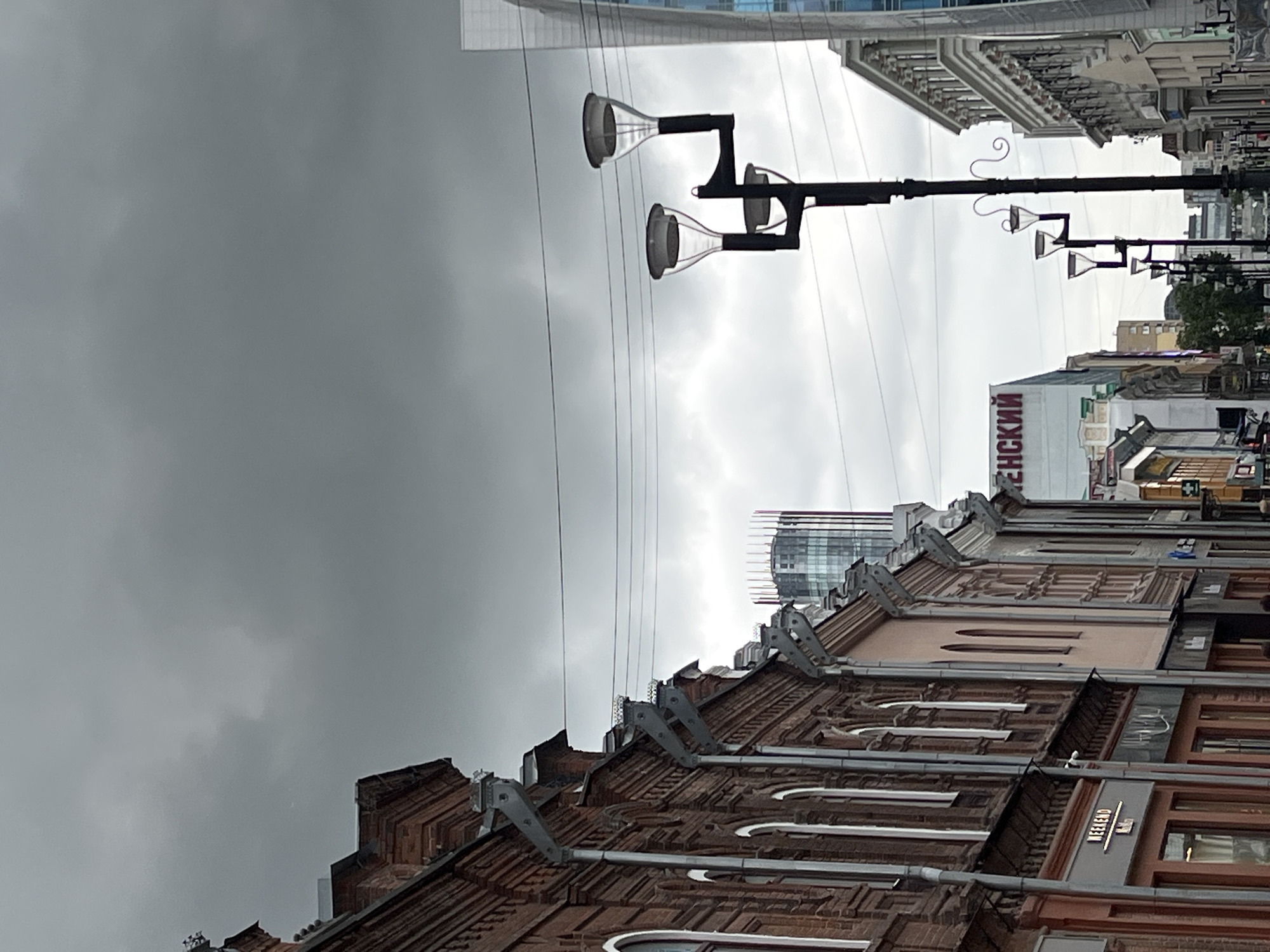The image depicts a street scene in Yekaterinburg, Russia, under a cloudy sky. On the left, a historic brick building with arched windows and ornate detailing lines the street. Further down, the facade of a modern building features large red lettering that reads "ЕНСКИЙ." Several lampposts, some with multiple lights, are visible along the street. In the background, a tall, modern skyscraper with a distinctive cylindrical design dominates the skyline. The street appears to be a commercial area, with storefronts visible on the ground floor of the older buildings. The overall atmosphere is overcast and urban.