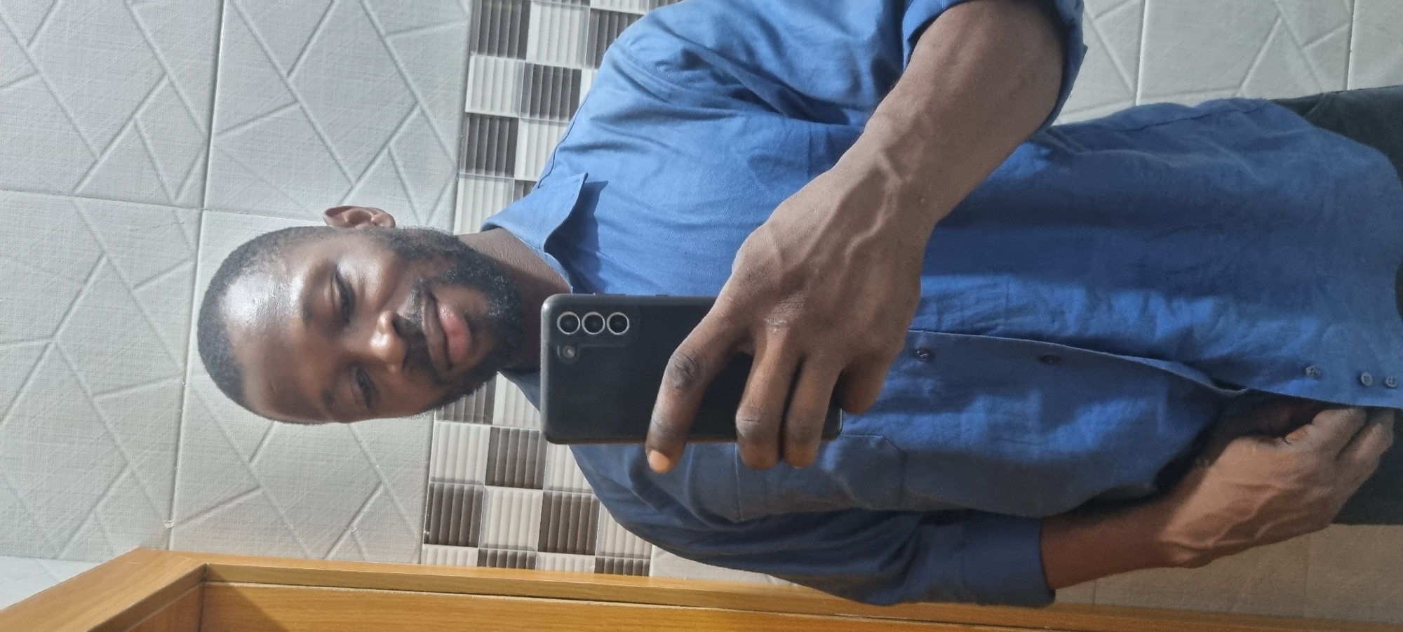 A man takes a selfie in what appears to be a bathroom. He has dark skin, short hair, and a beard. He is wearing a blue button-down shirt. He is holding a black smartphone in his right hand, likely to take the photo. The background consists of tiled walls, some patterned with geometric shapes and others with vertical stripes. A wooden frame is visible at the bottom of the image, possibly from a mirror or cabinet. The location is presumed to be Lekki, Nigeria. There is no further information available.