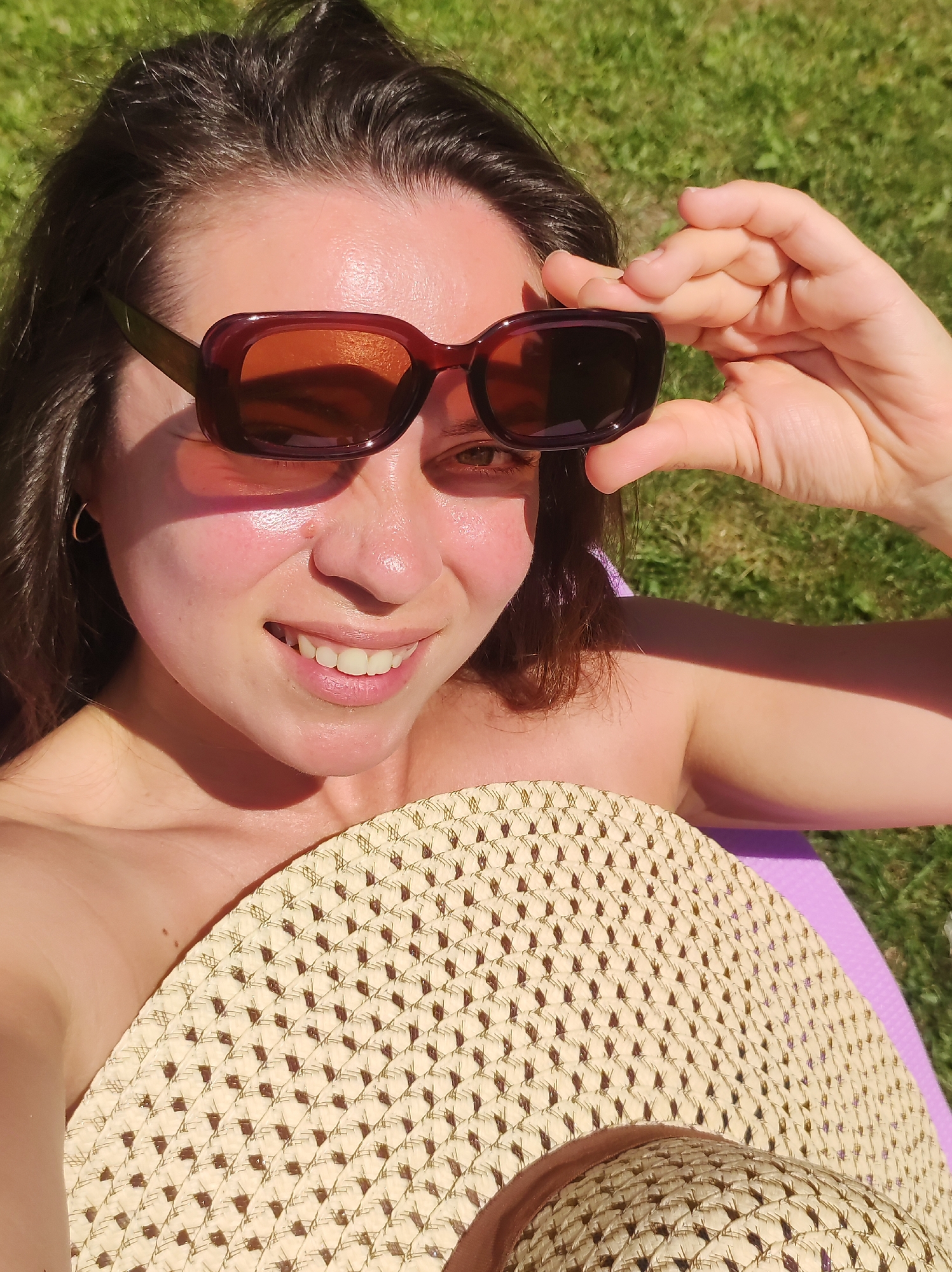 A young woman with dark hair and brown eyes is outdoors on a sunny day, looking at the camera while adjusting her sunglasses. She is wearing a straw hat. The background is a grassy area. The image does not provide information to determine a location in the city of Фоки, Russia.