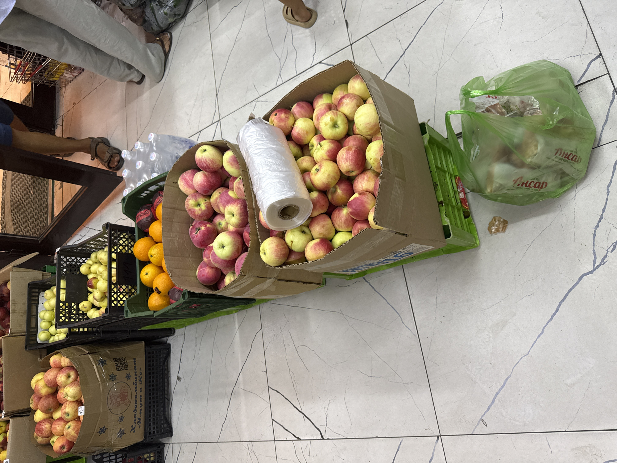 In Osh City, Kyrgyzstan, an outdoor market scene unfolds on a light-colored tiled floor with gray grout lines. Several cardboard boxes overflowing with fresh produce are arranged in rows. The boxes contain a variety of apples in shades of red and yellow, as well as oranges and what appear to be plums or other dark-skinned fruits. To the right of the fruit display, a person's legs are visible, wearing brown sandals and a light-colored skirt, indicating their presence in the scene. Near the center of the image, a roll of clear plastic wrap sits atop a box of apples. In the foreground, a translucent green plastic bag filled with items, possibly food or more produce, rests on the tiled floor. The background shows glimpses of a doorway, a shopping cart, and the legs of another individual wearing sandals and light-colored pants. The overall scene depicts a bustling marketplace environment.