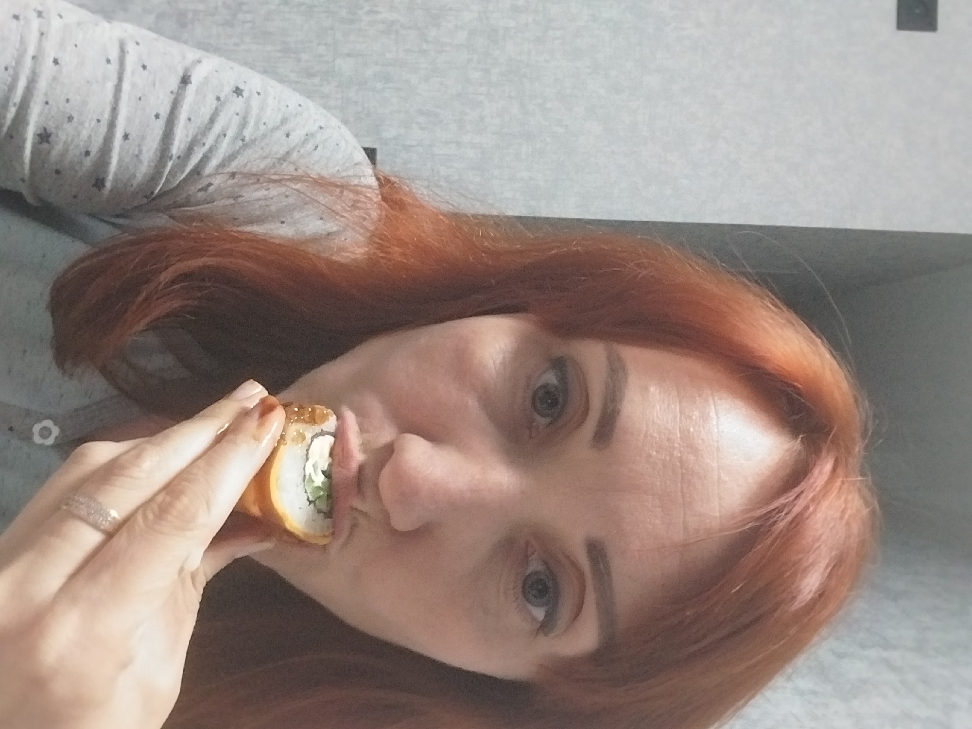A woman with reddish-brown hair and fair skin is shown from the chest up. She is wearing a light grey shirt with small dark star patterns. The woman is bringing a sushi roll to her mouth with her right hand. Her fingers are visible, and she is wearing a ring on her right ring finger. The background appears to be an indoor setting with a textured, light-colored wall. There is no discernible indication of the specific location within the city of Dnipro, Ukraine.