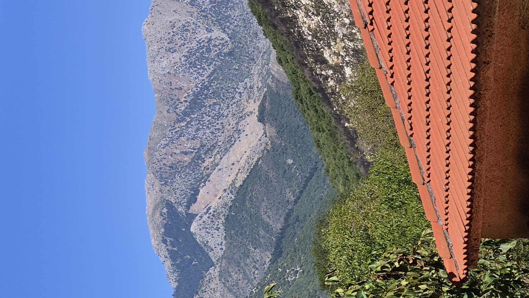 The image depicts a scenic mountainous landscape under a clear blue sky. In the foreground, a portion of a building with a terracotta tiled roof is visible. The tiles are uniformly red and exhibit a wavy pattern along their edges. The building's wall, visible beneath the roof, is textured and appears to be made of a rough material.
Behind the building, a lush green foliage is present, with trees and bushes. Beyond the immediate greenery, rolling hills covered in dense vegetation stretch into the distance. The middle ground is dominated by layers of mountains, with varying shades of green indicating forested areas and lighter, rocky slopes.
The background features a prominent mountain range with rugged, rocky terrain. The upper slopes of these mountains are largely bare, with patches of scrub and lighter-colored rock formations. The overall impression is of a natural, undeveloped environment, possibly in a rural or mountainous region. There are no people or animals visible in the image. The lighting suggests it is daytime, with the sun casting some shadows but illuminating the scene clearly.