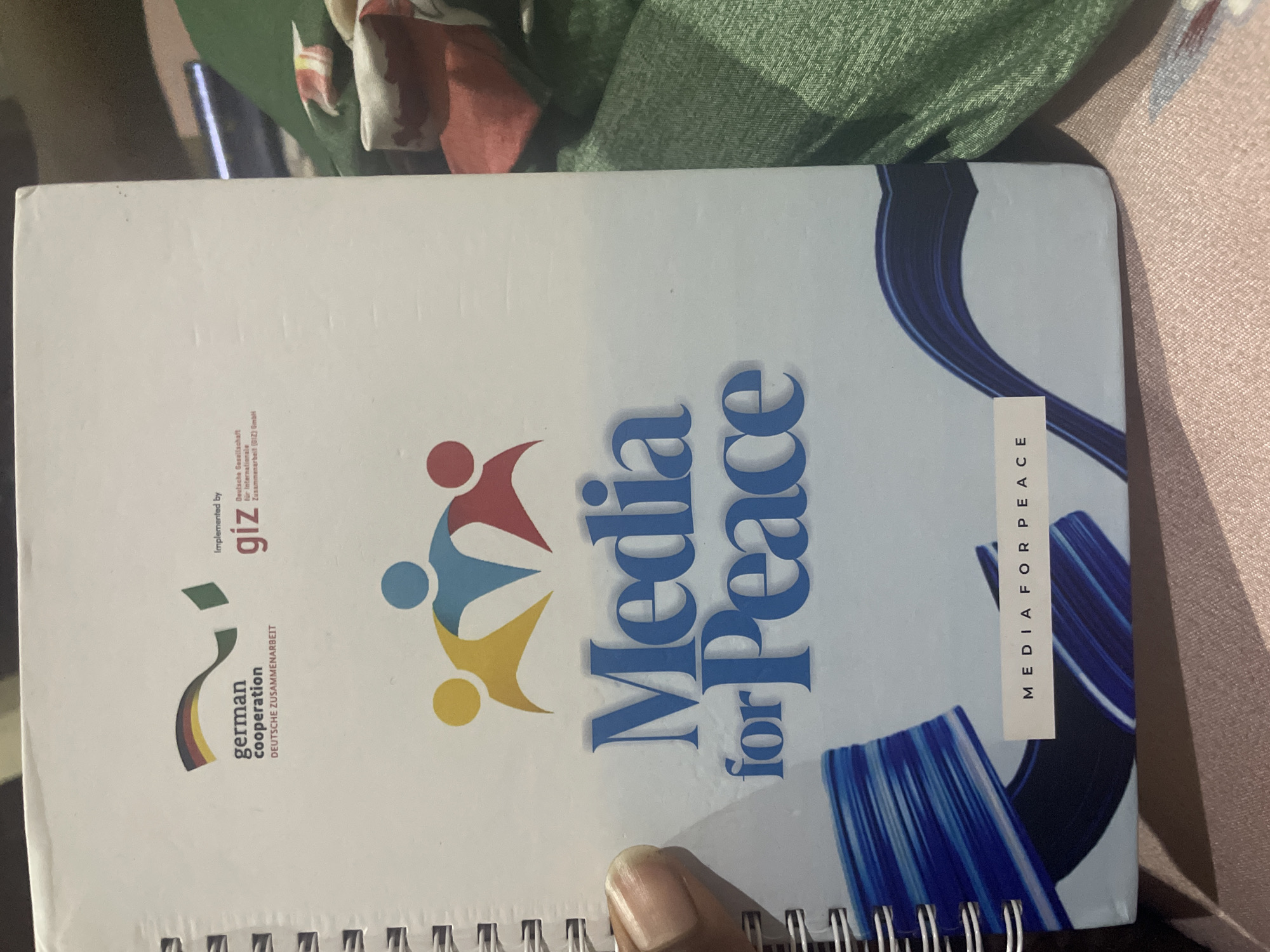 The image shows the cover of a notebook or binder with the title "Media for Peace" prominently displayed in large blue lettering. The cover features several logos and graphics, including the German flag colors forming a ribbon shape, the text "german cooperation DEUTSCHE ZUSAMMENARBEIT," and a logo for "giz" with "Implemented by Deutsche Gesellschaft für Internationale Zusammenarbeit (GIZ) GmbH." There is also a graphic of three figures linked together in yellow, blue, and red. A small white label at the bottom reads "MEDIA FOR PEACE." The notebook is lying on a patterned surface, possibly a tablecloth or fabric, and a green fabric is visible in the background. The scene is dimly lit, and the notebook is slightly angled. There is no information in the image to suggest the location is Jalingo, Nigeria.