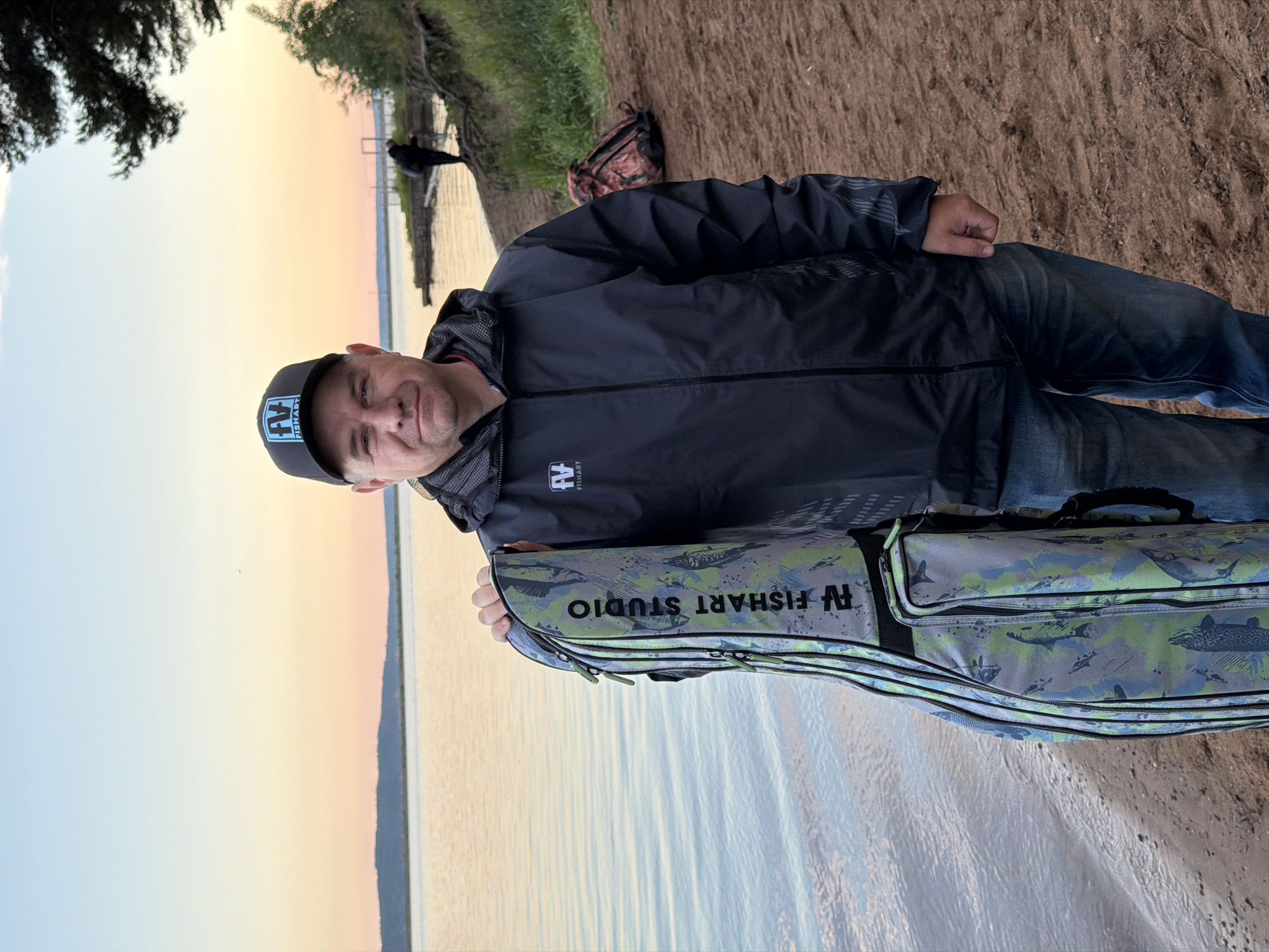 A man stands on a sandy or dirt bank beside a body of water, likely a lake or river, in Chaikovsky, Russia, during sunrise or sunset. He is depicted from the waist up, looking at the camera with a gentle smile. He wears a dark, possibly black or charcoal gray, jacket with patterned sleeves, blue jeans, and a dark baseball cap that features a logo with the text "FV FISHART".
The man is holding a long, camouflage-patterned bag, presumably a fishing rod case, which also displays the text "FV FISHART STUDIO". The bag has several zippered compartments.
The background to the left (relative to the man) shows a calm expanse of water reflecting the sky's soft colors, transitioning from light blue above to warm orange and yellow closer to the horizon, indicative of the golden hour. A distant, dark landmass or structure is visible across the water. To the right and behind the man, the sandy ground continues, and a red and camouflage-patterned backpack is visible. Further in the distance, a dark bird perches on a wooden structure extending into the water. Dark green tree foliage is visible in the upper corners, framing the scene and suggesting a natural, possibly wooded, environment. The overall mood is peaceful and serene.