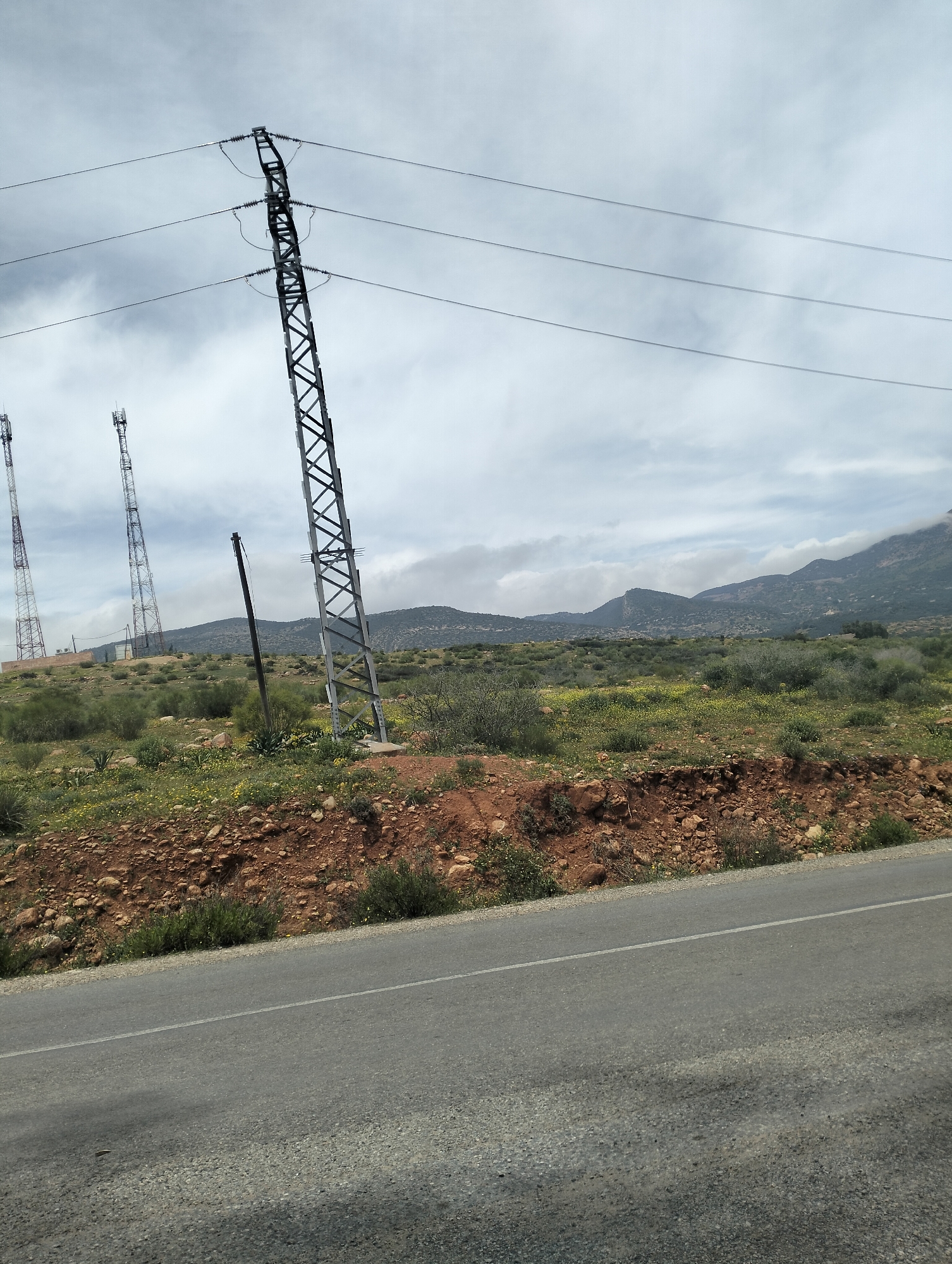 The image shows a roadside scene in what appears to be Ouaouizert, Morocco. An asphalt road with a white lane divider is visible in the foreground. Beyond the road is a low, eroded embankment of reddish-brown earth with sparse green vegetation and small rocks. Further back is a landscape of scrubby vegetation interspersed with yellow wildflowers, leading to a mountainous horizon partly obscured by a cloudy sky. An electrical transmission tower stands prominently to the left of the frame, with power lines extending across the top portion of the image. In the distance to the left are two additional telecom towers, likely transmitting cellular signals. A dark, thin utility pole is also visible in the middle ground.