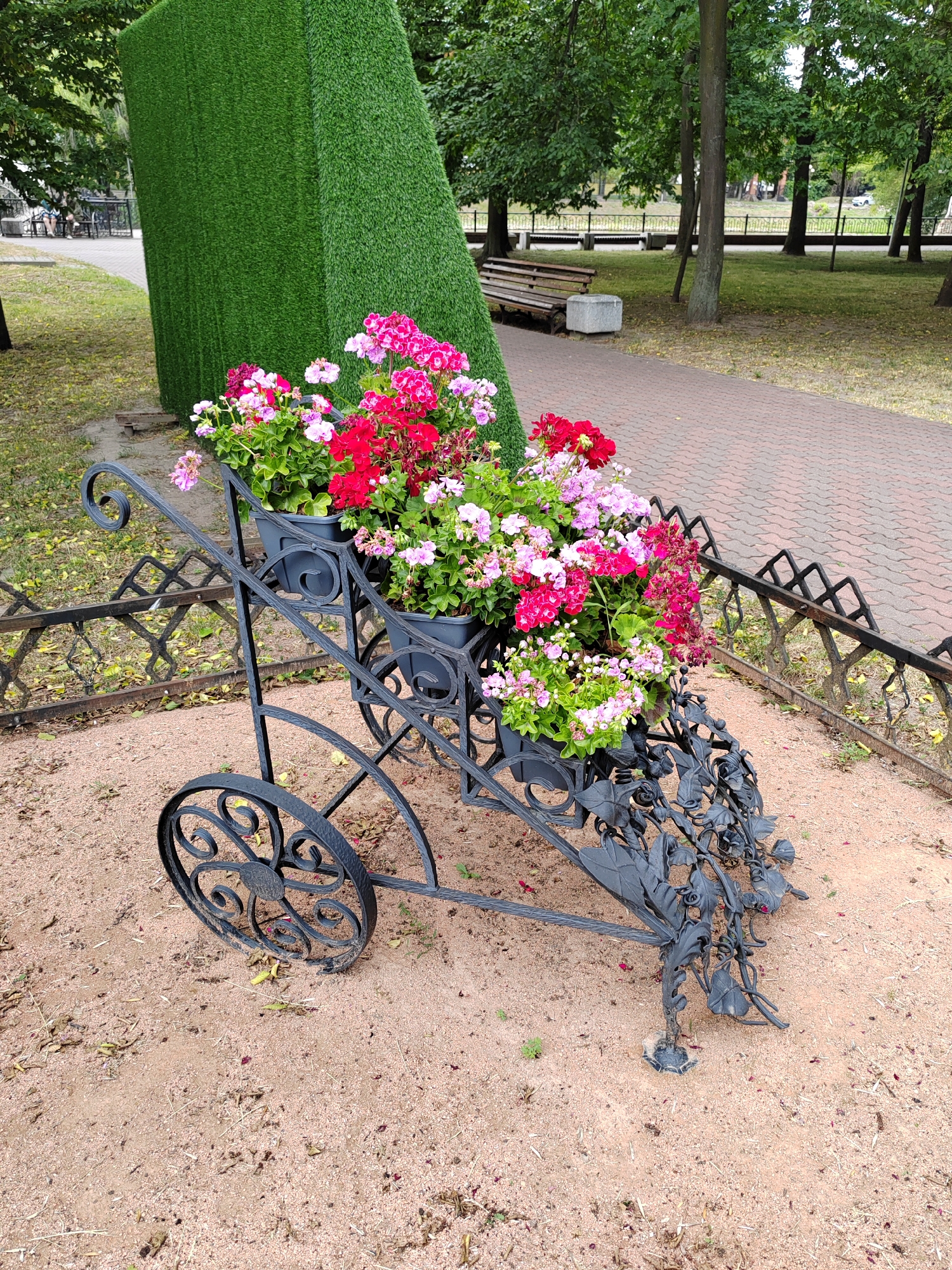 In Brest, Belarus, an ornate black wrought-iron cart serves as a planter, overflowing with vibrant pink and red geraniums. The cart, with decorative wheels and scrollwork, is positioned on a sandy path in a park-like setting. Behind the cart, a tall, angular structure covered in artificial green turf stands prominently. In the background, a paved walkway curves through a grassy area dotted with mature trees. A wooden bench with a stone base is visible near the trees, and a decorative black metal fence runs along the edge of the path. The scene suggests a manicured urban green space.