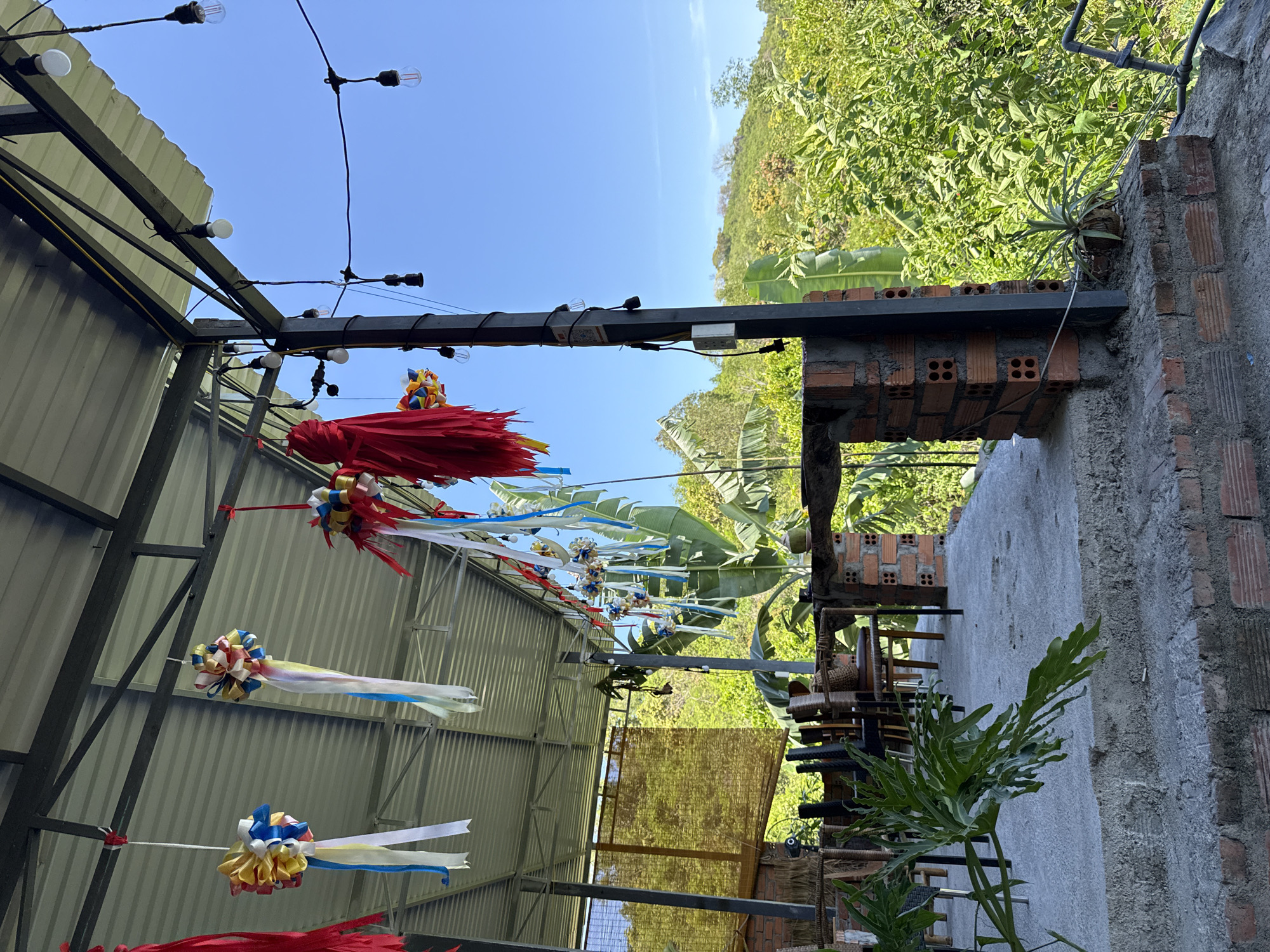 This image depicts an outdoor covered seating area with a corrugated metal roof, supported by metal beams. Decorative ribbons in red, yellow, blue, and white hang from the roof. Strings of bare light bulbs are also suspended from the beams. The seating area appears to be constructed with a concrete floor and brick supports. Various chairs and tables are visible. Beyond the seating area, lush green vegetation and trees dominate the landscape, with a hilly terrain in the background. The sky is a clear, bright blue. There are no people visible in the image. The scene is likely a restaurant or cafe in a tropical or subtropical location.