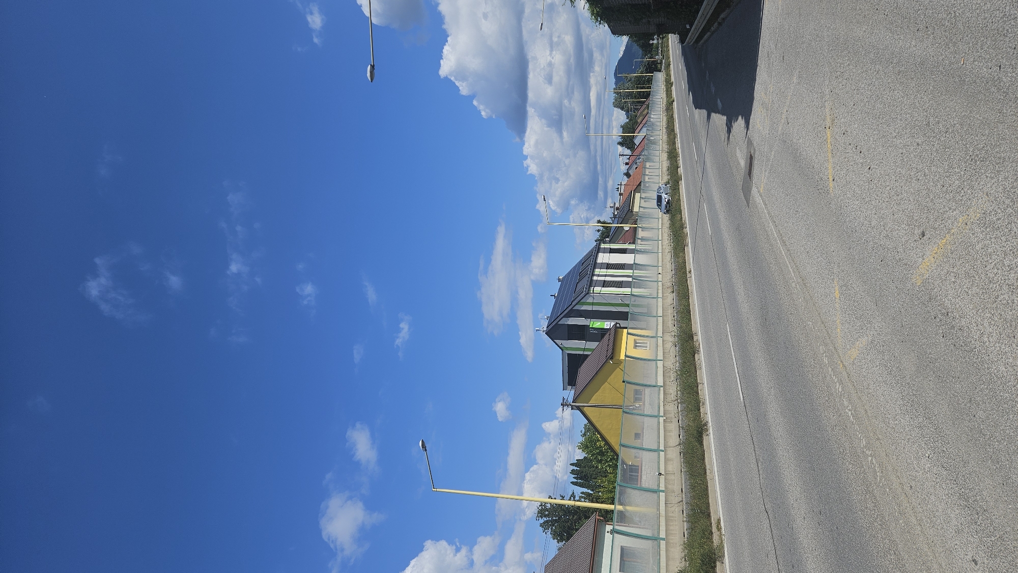 The image depicts a street scene in Prešov, Slovakia, on a bright, sunny day with a clear blue sky dotted with white clouds. A paved road runs through the foreground, with a dividing line visible. To the left of the road, there is a concrete barrier topped with a green and transparent fence, separating the road from a row of houses.
The houses exhibit various architectural styles and colors. One prominent building in the middle features a modern design with a black and white striped facade and a dark roof covered in solar panels. Adjacent to it is a bright yellow house with a brown roof. Further down the row, other houses with orange and red tiled roofs are visible.
Streetlights are positioned along the road and the houses. A silver car is parked or driving on the road in the distance. The background shows trees and a gently sloping hill with a mountain peak obscured by clouds. The overall environment suggests a suburban or semi-urban setting in Prešov.